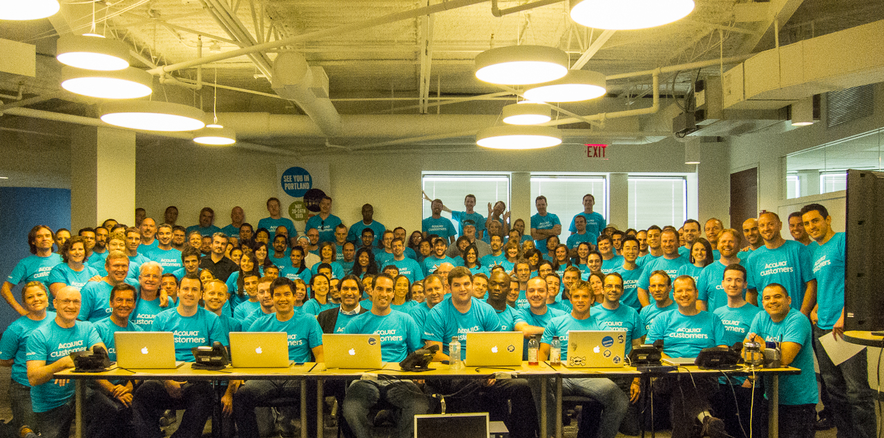 A large group of Acquia employees in matching blue shirts gathers in an office to celebrate Customer Support Day in July.