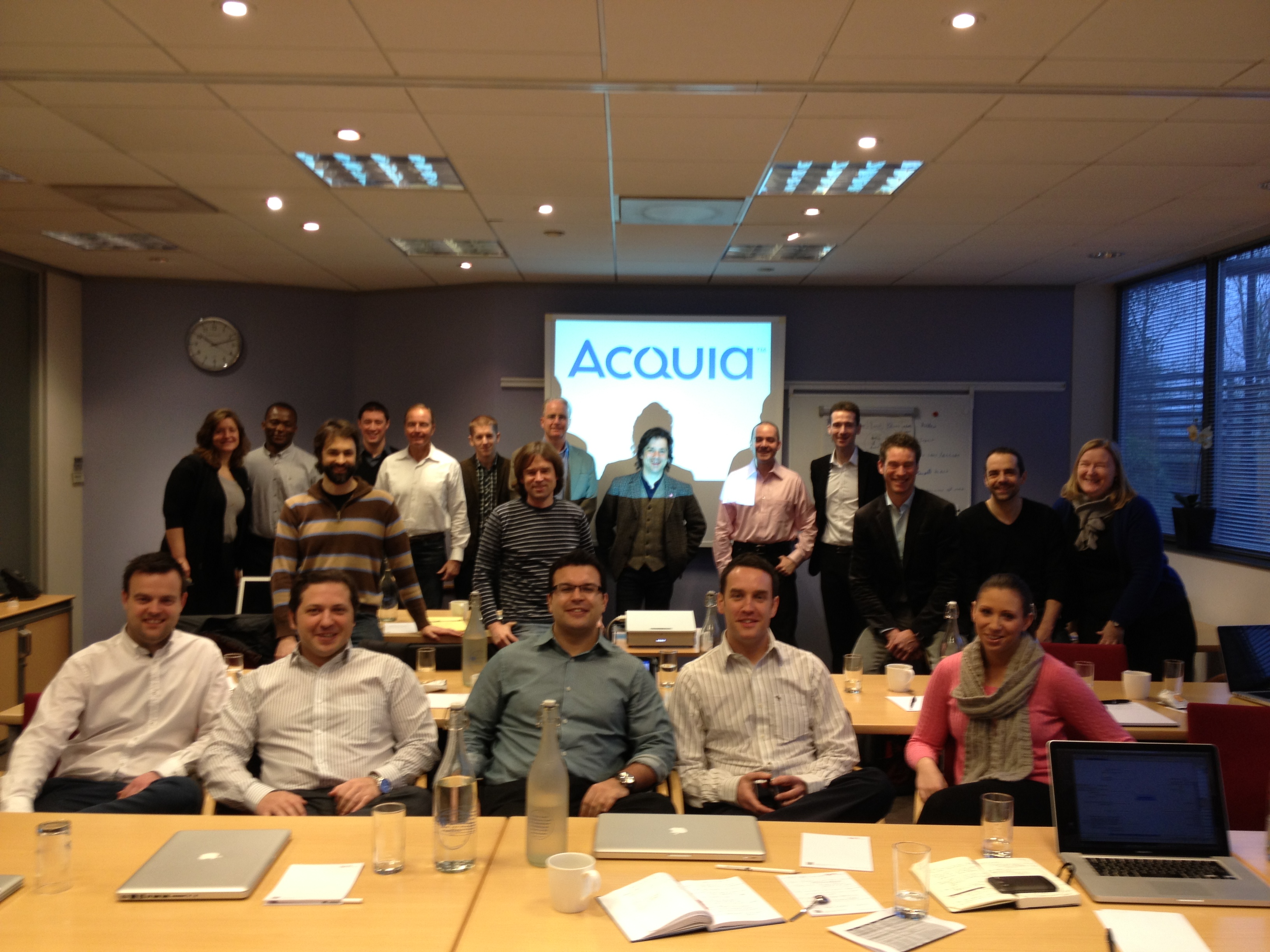 A group of people posing in a conference room with an "Acquia" presentation displayed on the screen behind them.