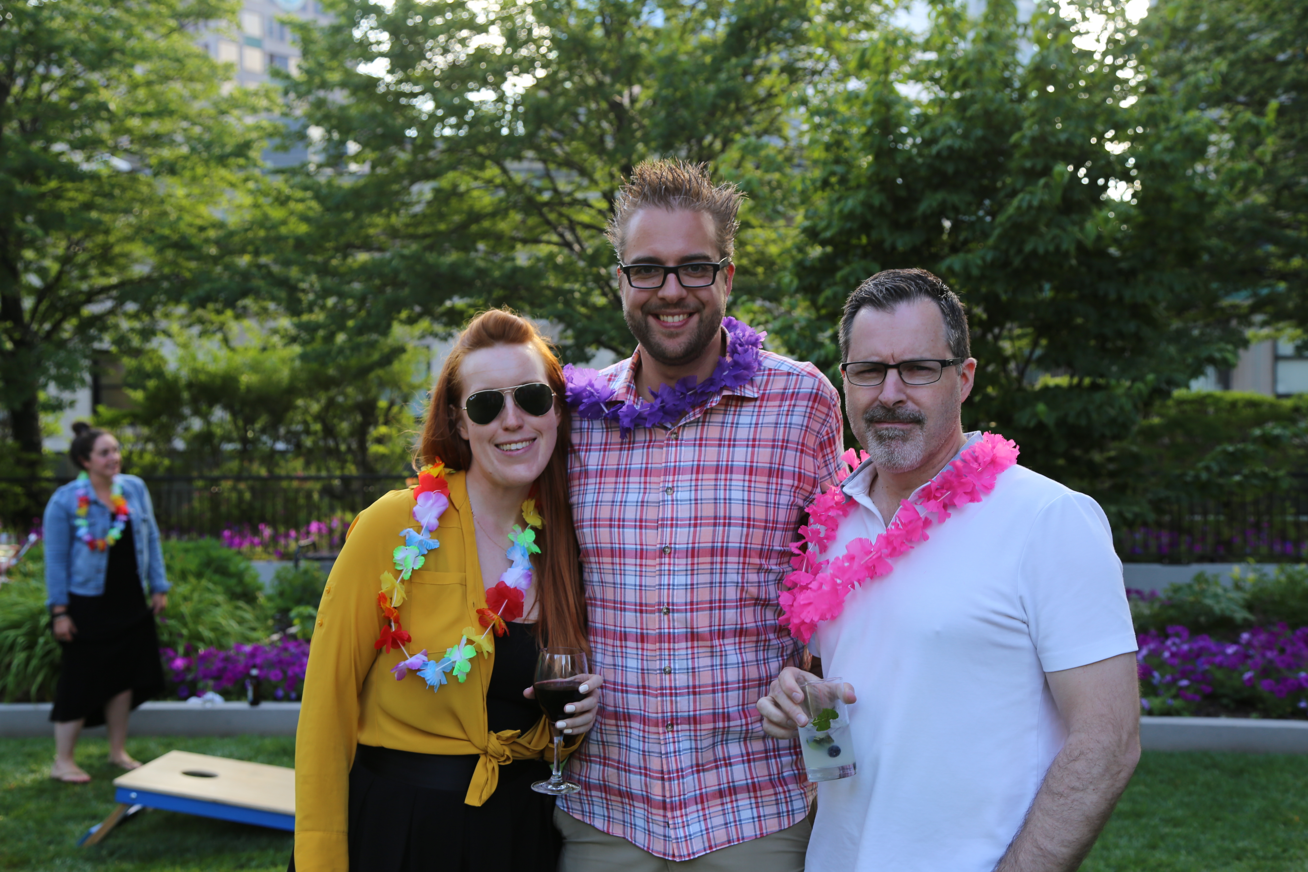 Three people wearing colorful leis smile together outdoors, holding drinks.