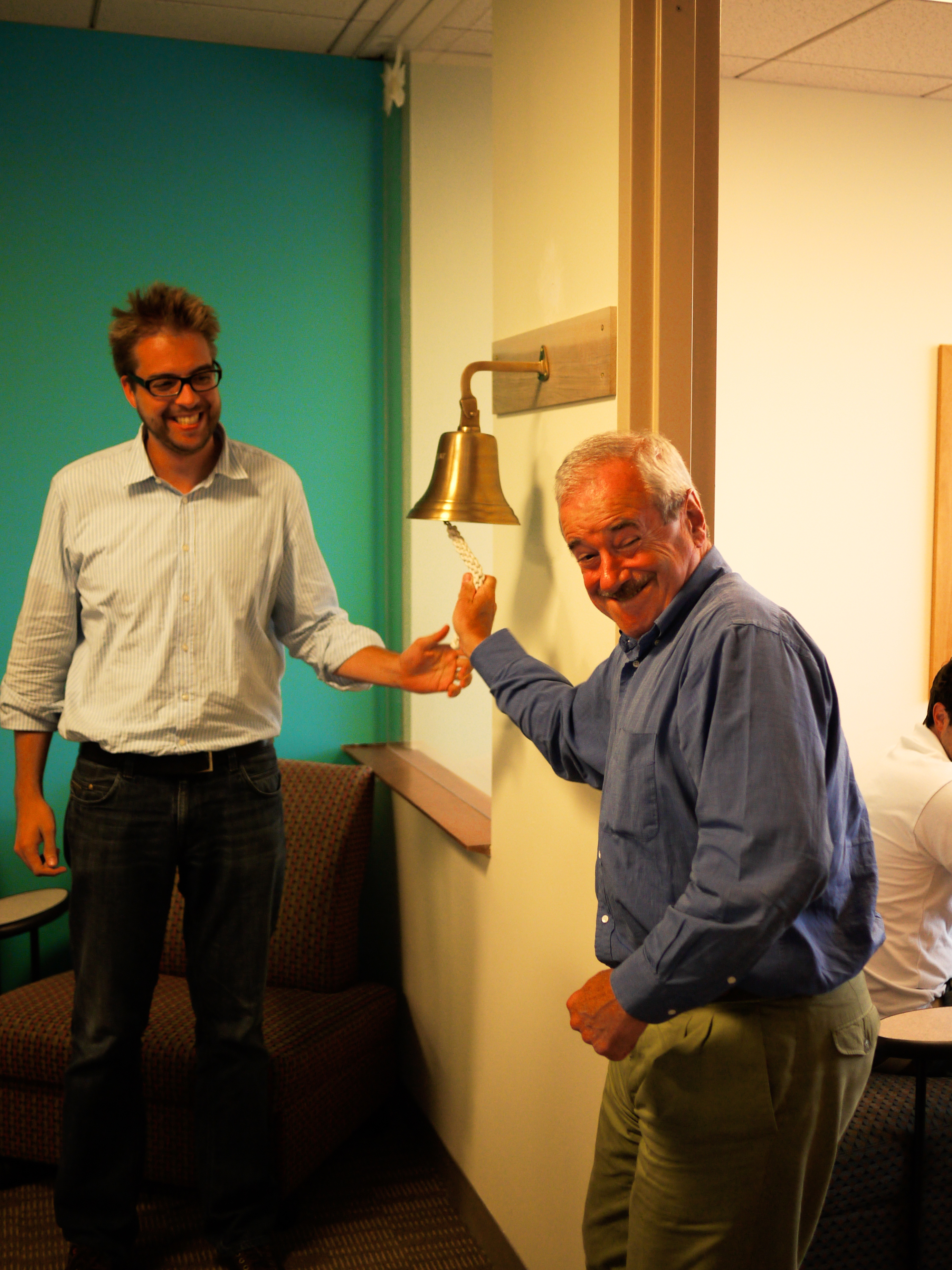 An older man rings a wall-mounted bell while a younger man watches and smiles.
