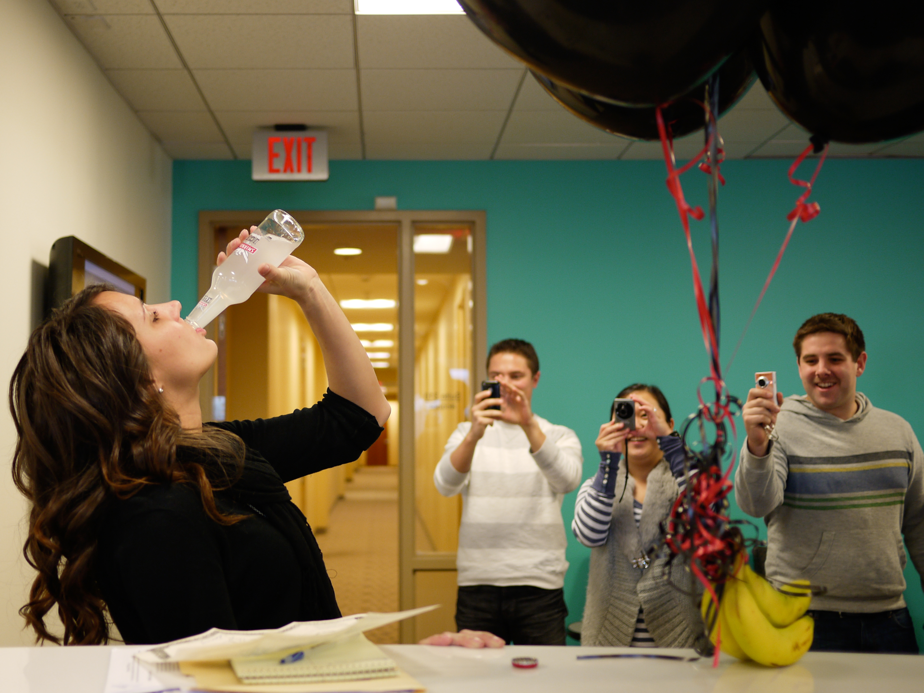 A woman drinks from a bottle while three people take photos and smile in a decorated office.