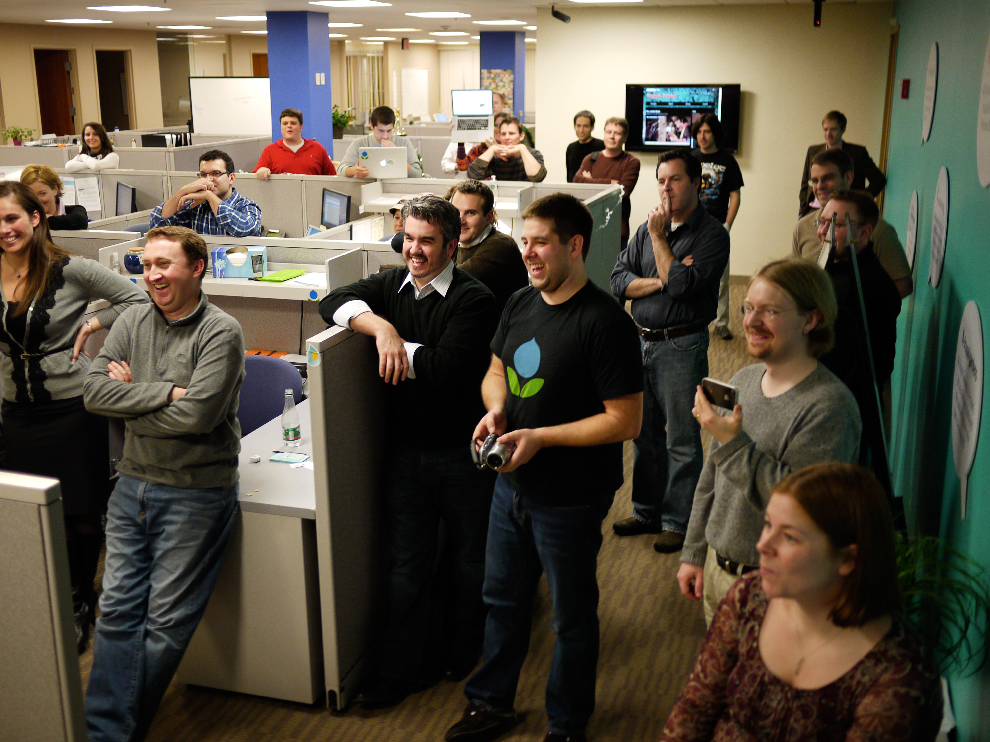 Coworkers in an office gather, smiling and laughing while watching something off-camera.
