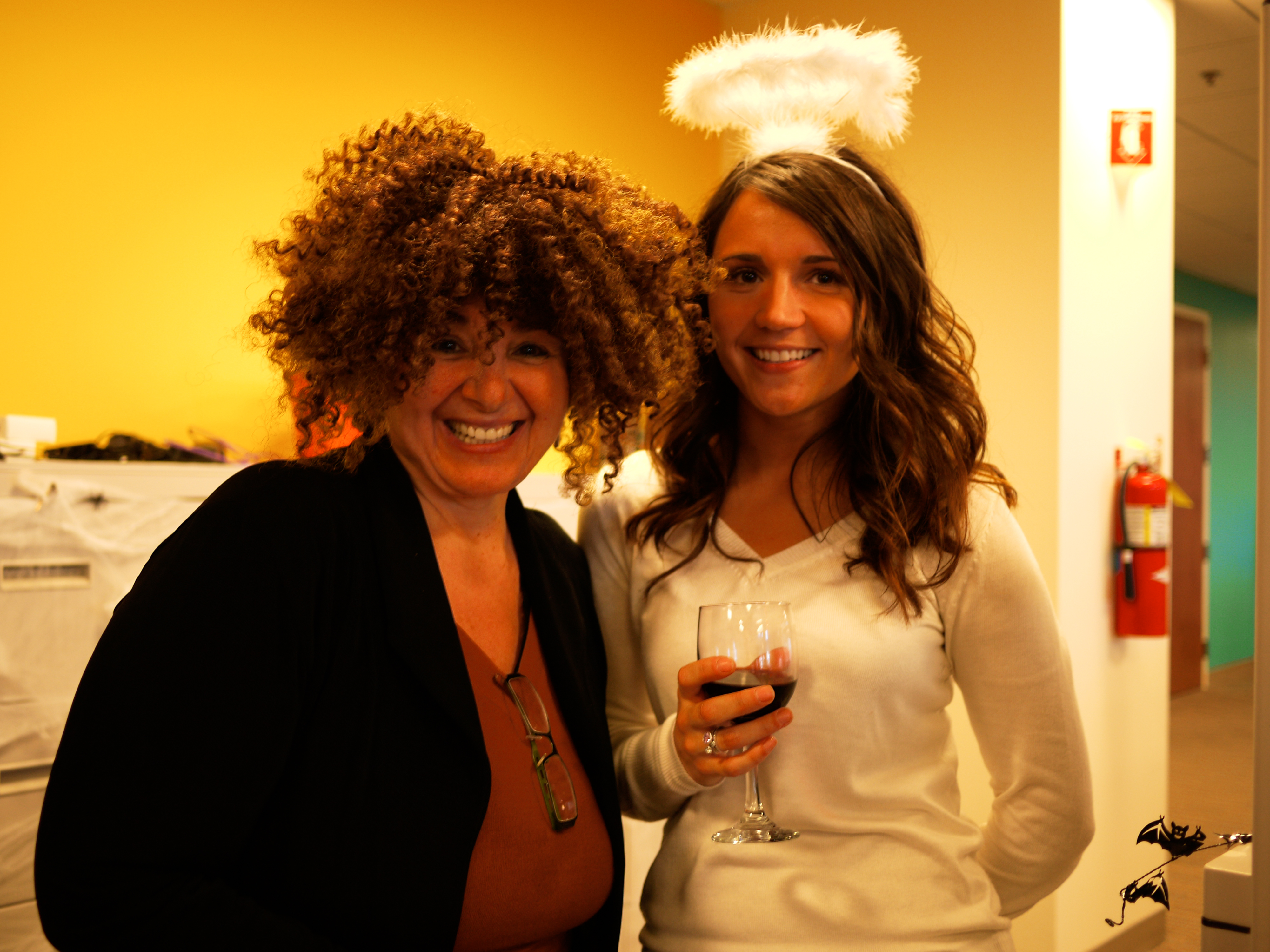 Two people in Halloween costumes smile at the camera; one wears a curly wig, the other an angel halo.