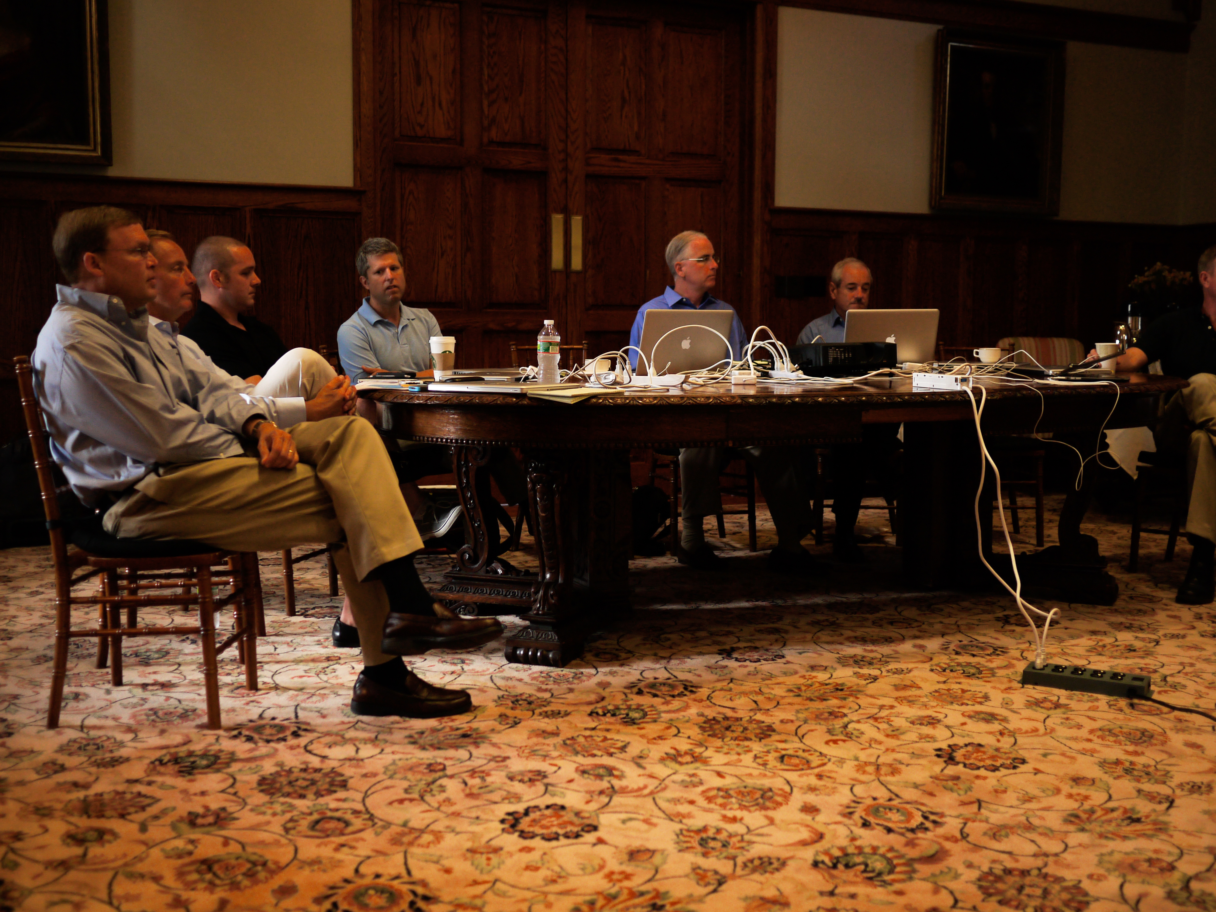 A group of people sit around a table with laptops and cables during a leadership offsite meeting.