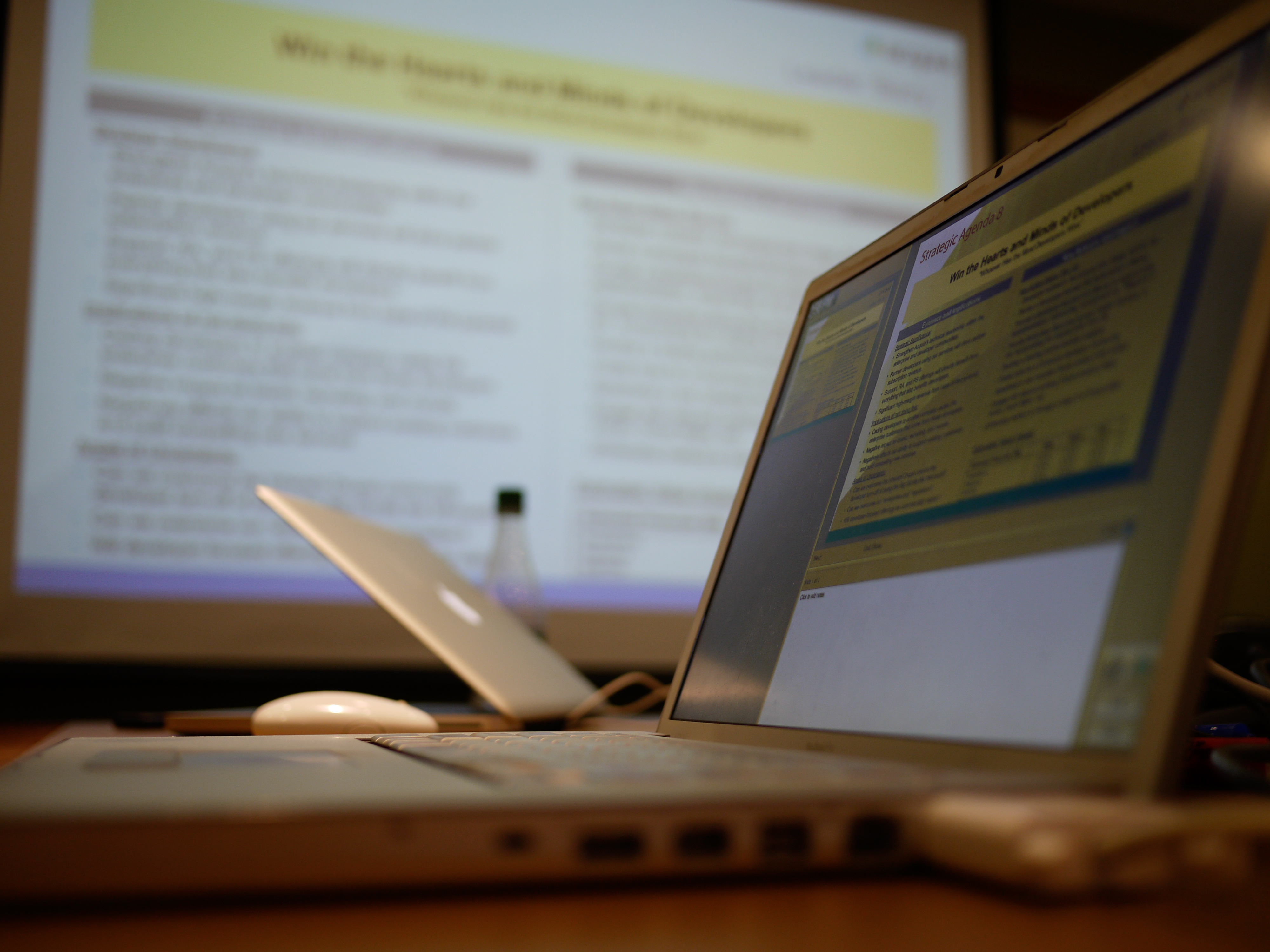 Two laptops on a table display presentation slides, with a projector screen showing similar content in the background.