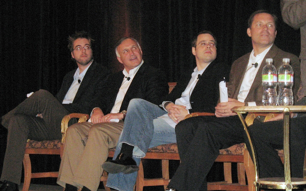 Four men on a panel discussion sit with microphones, listening attentively to someone off-camera.