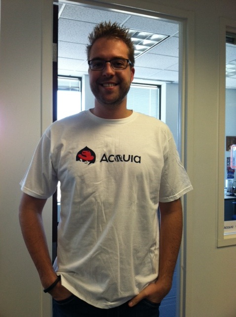 A person wearing a white Acquia T-shirt with a red Drupal logo, smiling indoors.