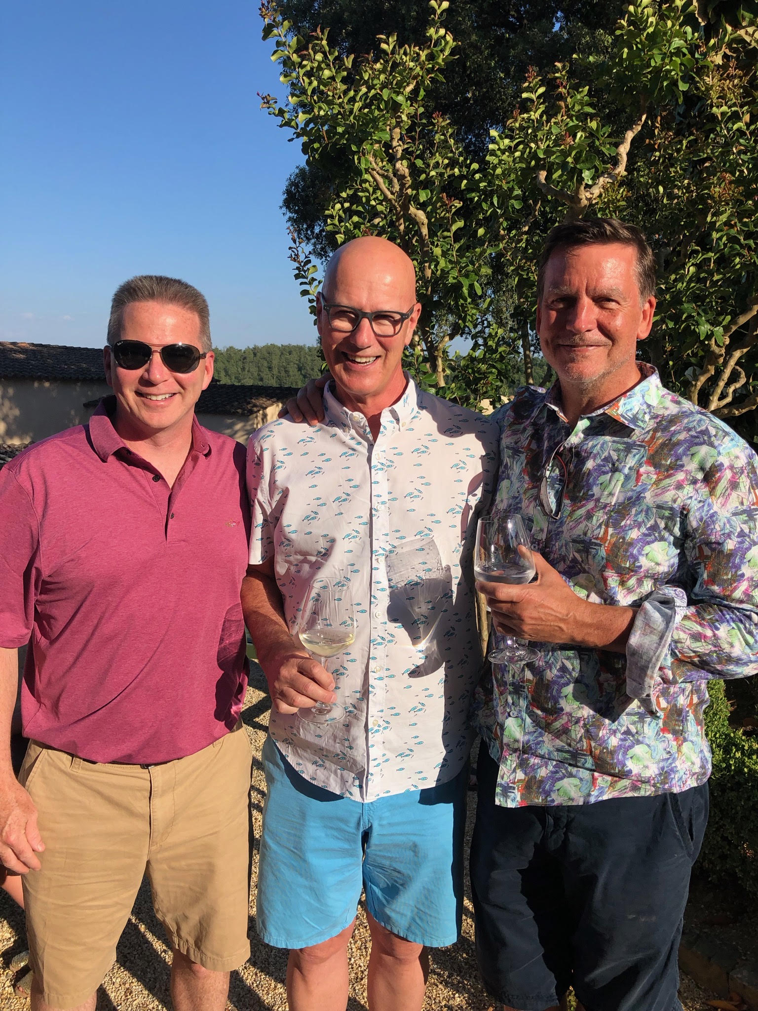 Three men stand together outdoors, smiling and holding wine glasses.