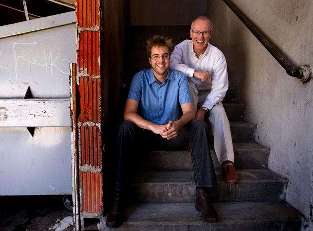 Two men sitting on outdoor concrete stairs, both smiling at the camera.