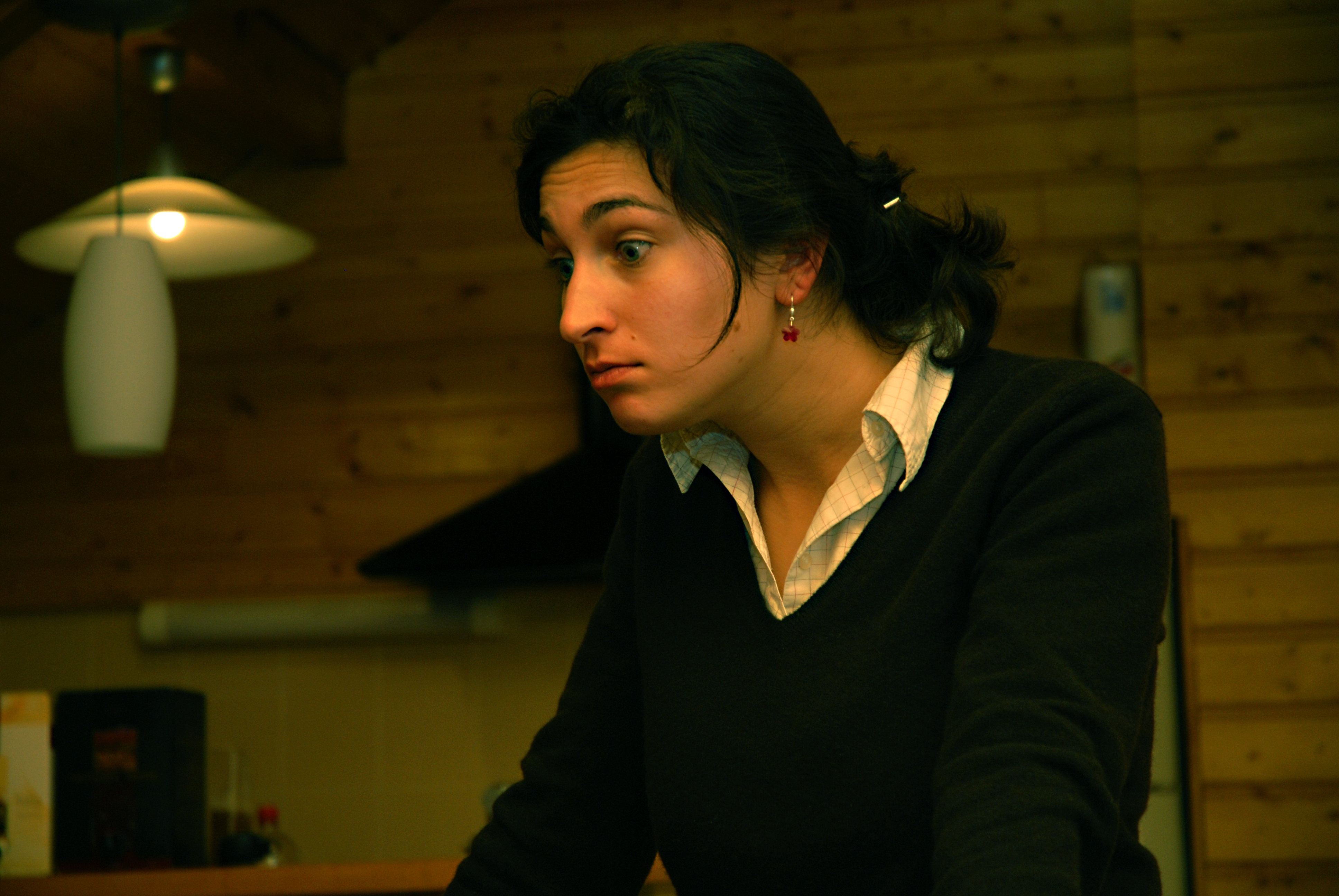A woman with dark hair and red earrings leans forward, looking intently at something with a focused expression.