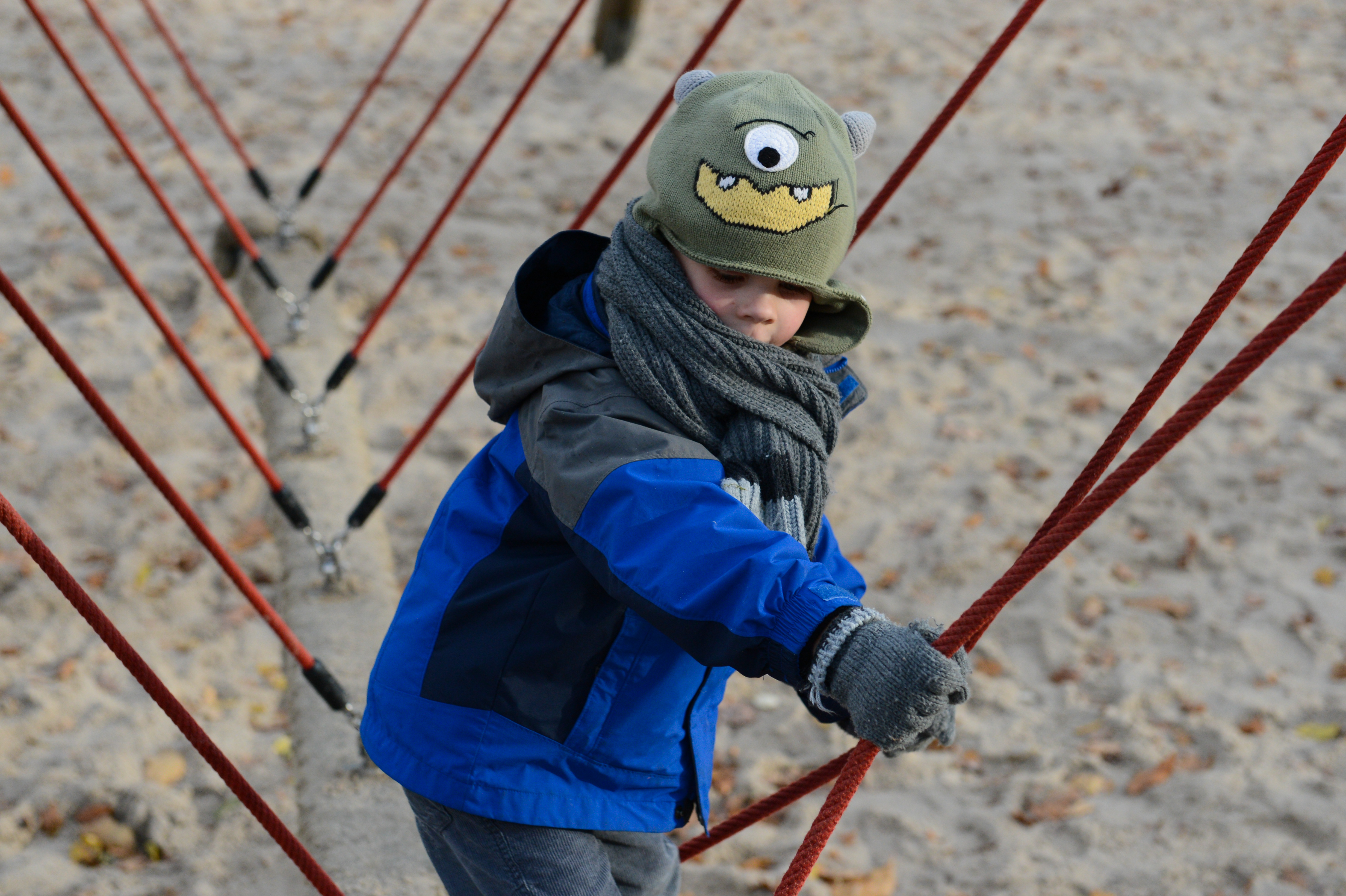 A child in a blue jacket and monster hat climbs a rope structure at a sandy playground.