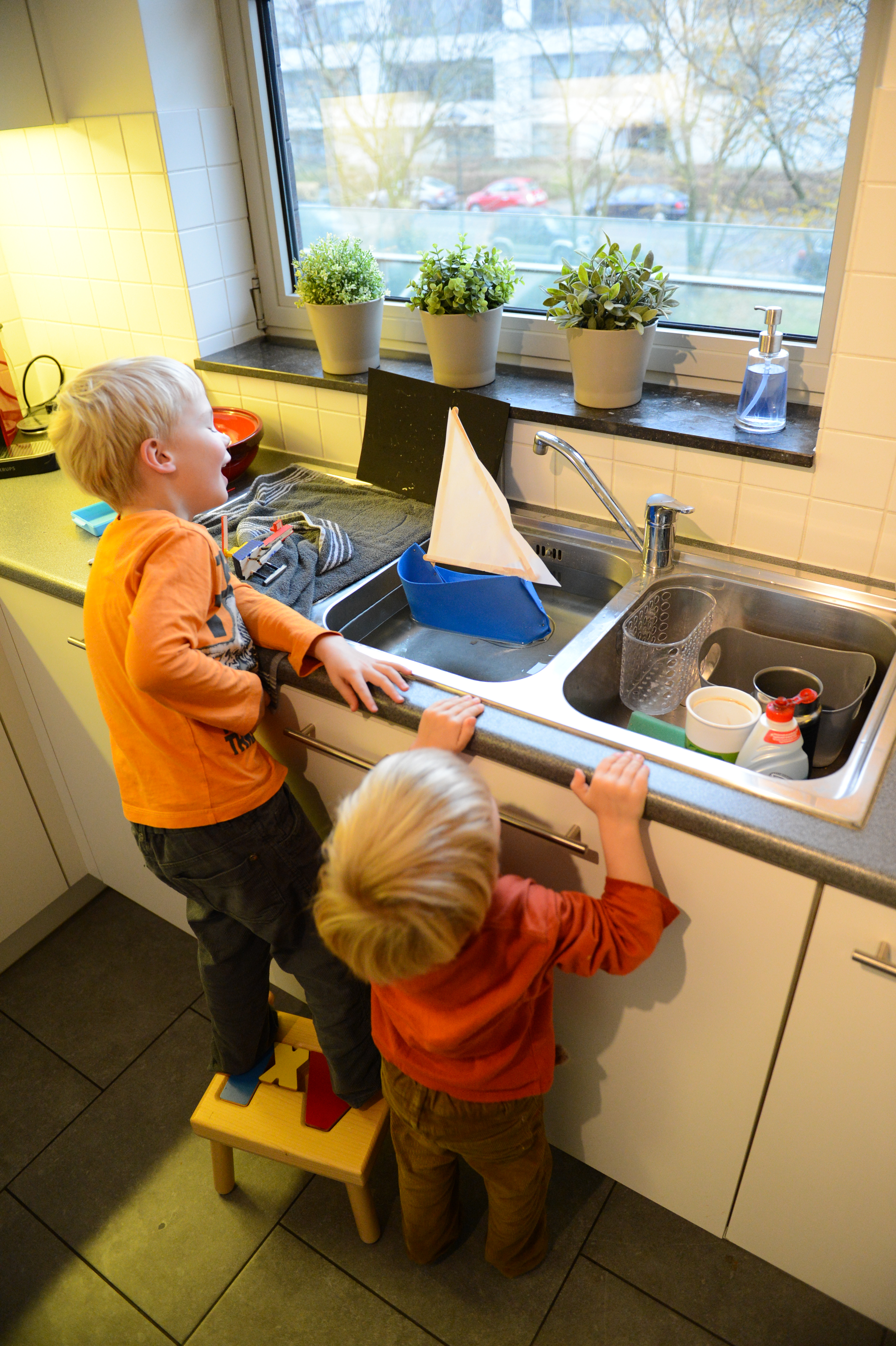 Two children in an orange outfit watch a small homemade sailboat floating in a kitchen sink.