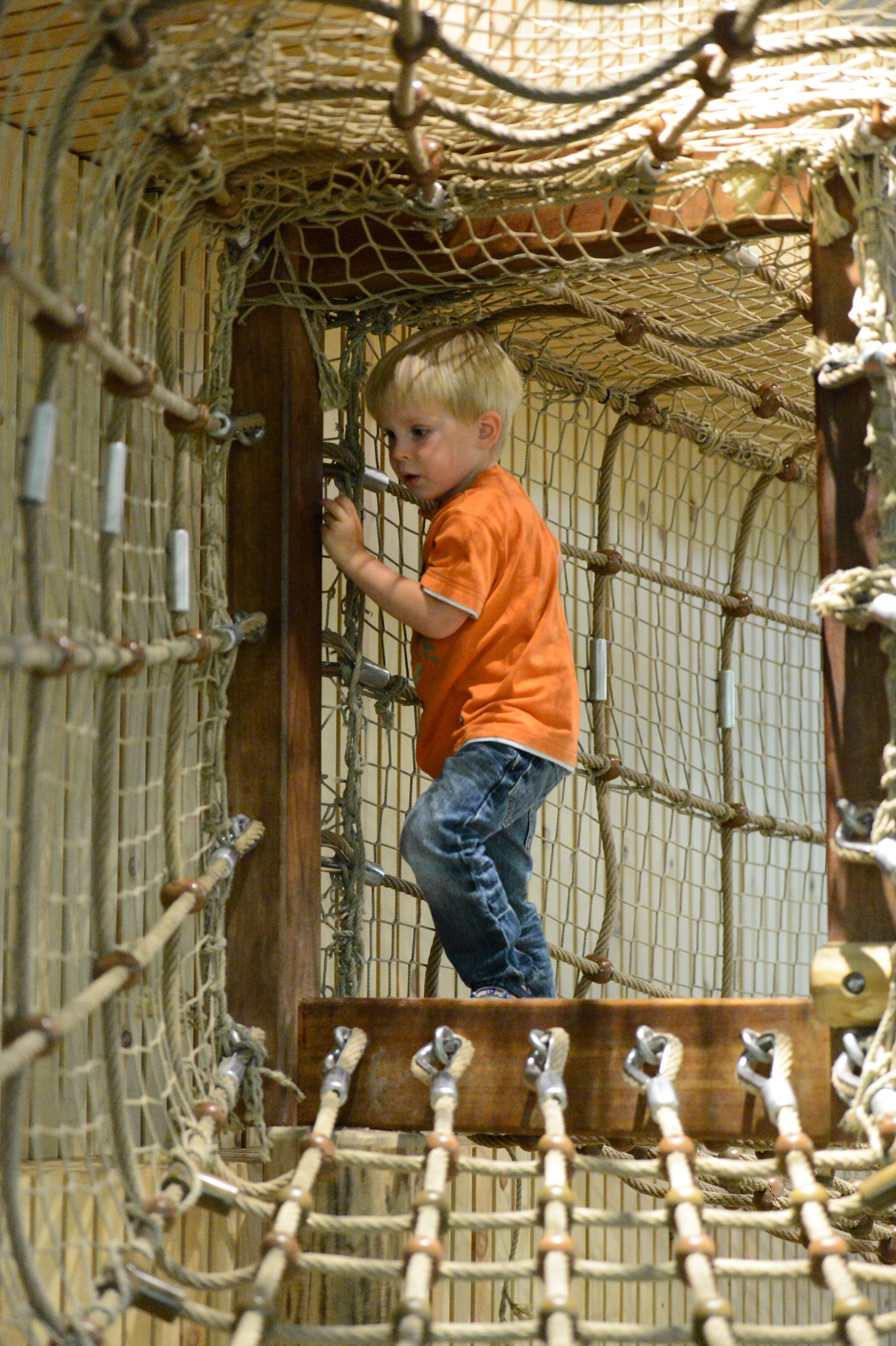 A young child in an orange shirt climbs through a rope tunnel at a zoo play area.