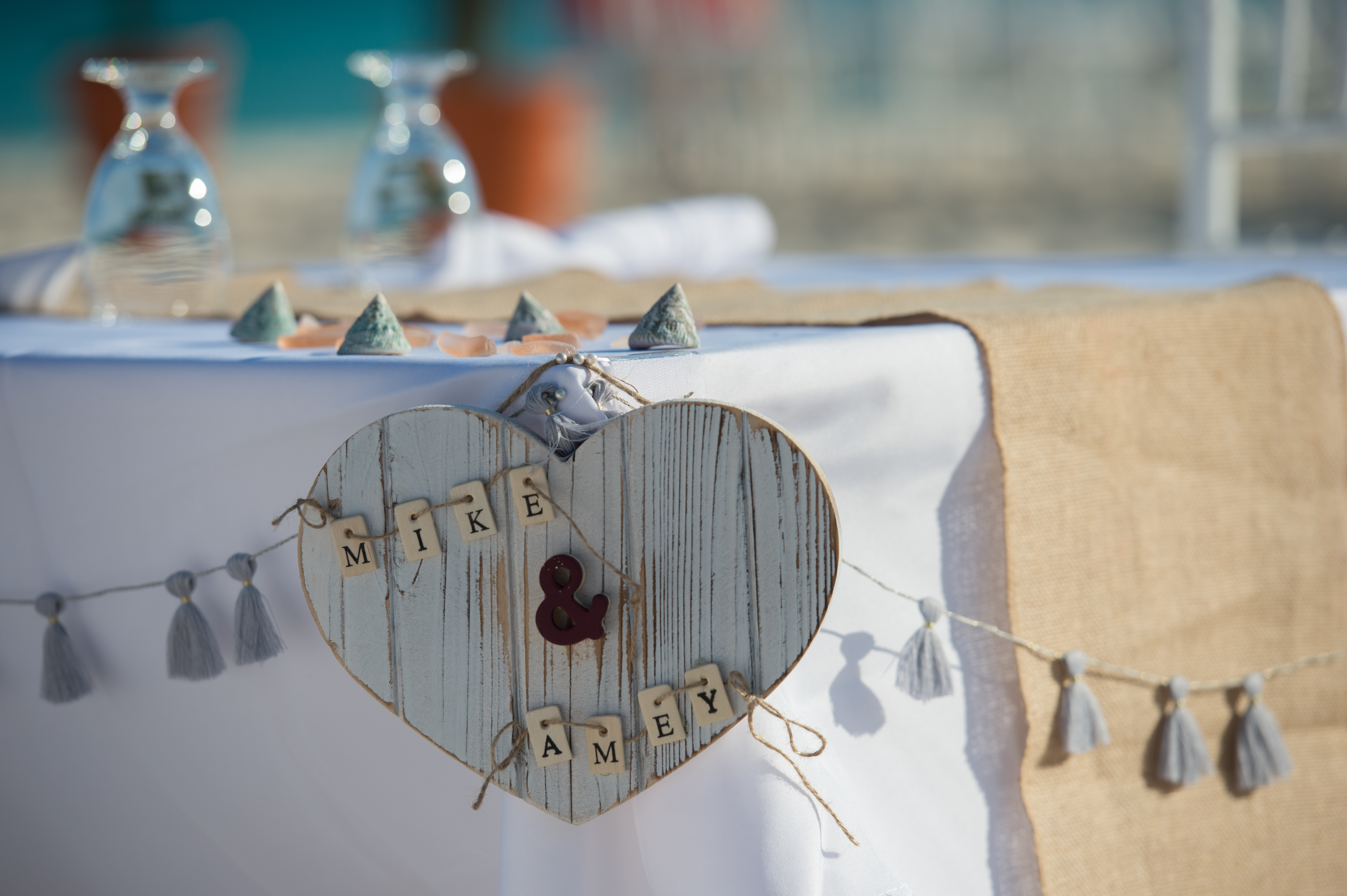 A heart-shaped wooden sign with "Mike &amp; Amey" hangs on a decorated wedding table with tassel garland.