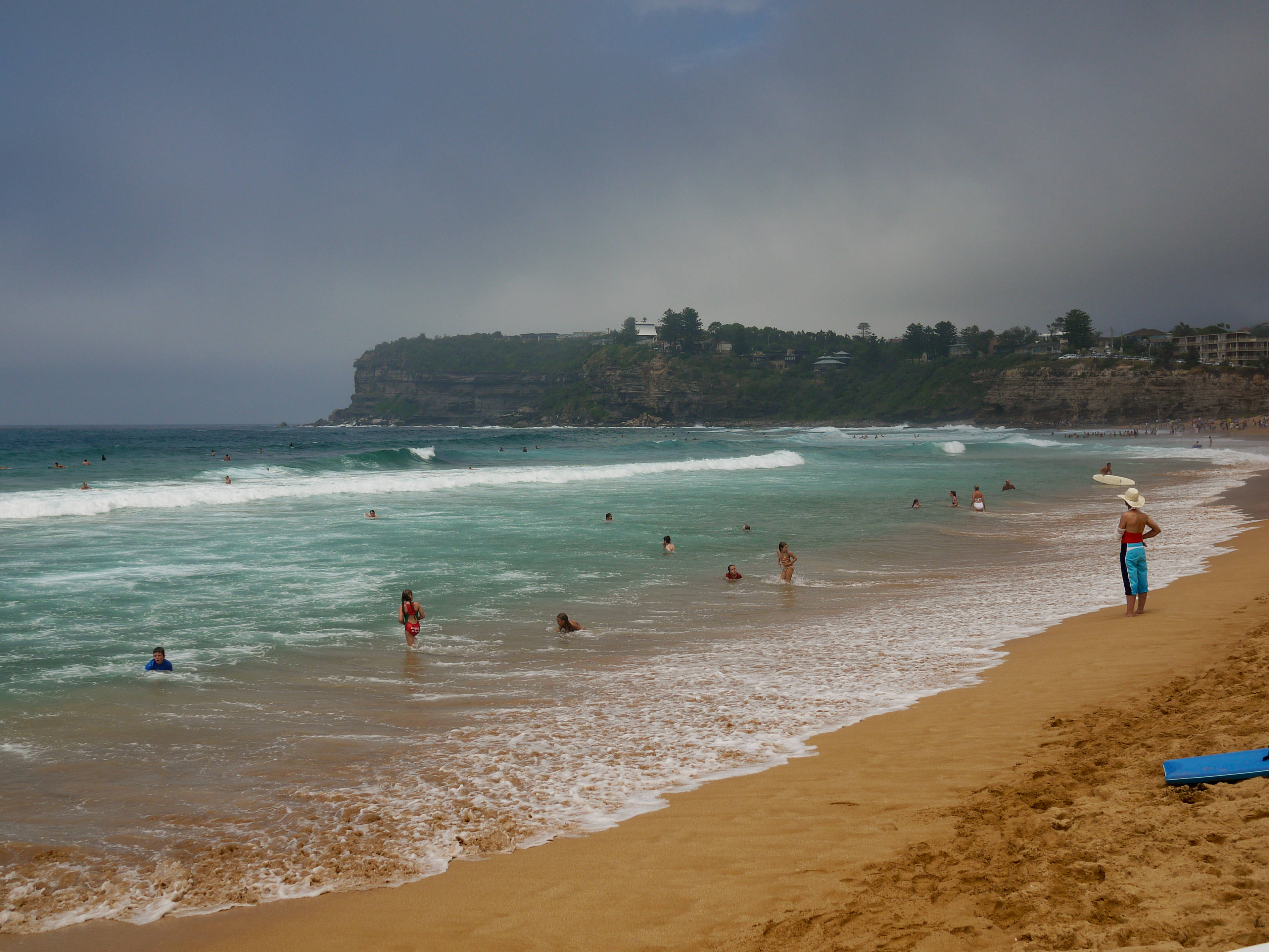 People swimming and wading in the ocean at Avalon Beach, Sydney, with a surfer standing on the shore.