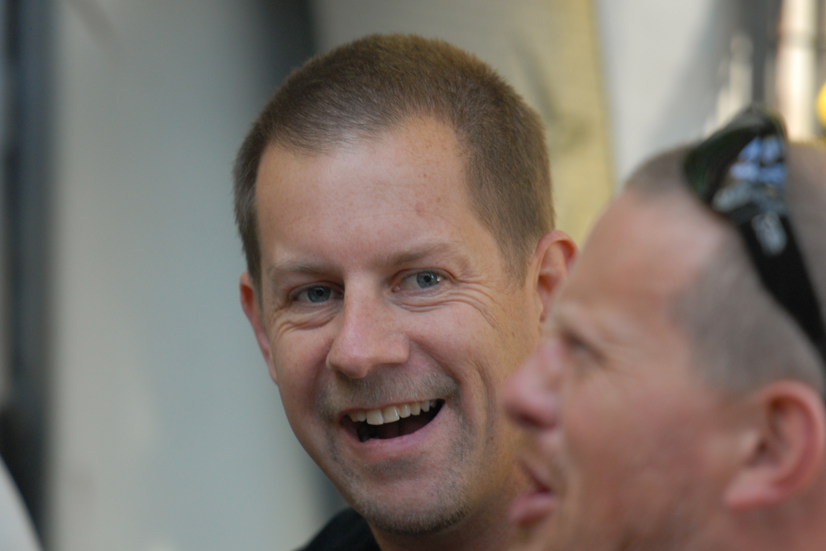 Two men at a party, one smiling and the other talking while wearing sunglasses on his head.