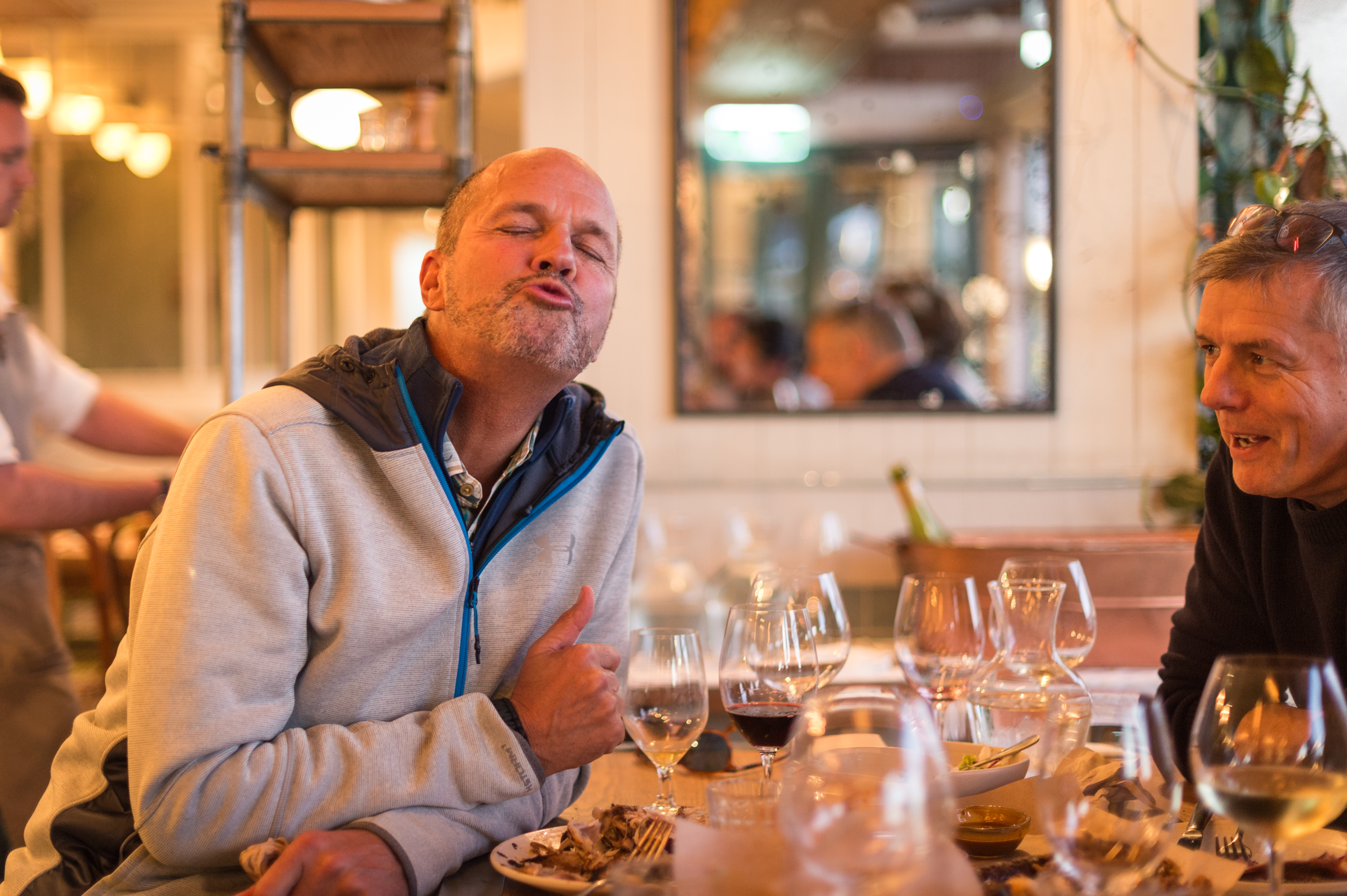 A man in a gray jacket puckers his lips playfully while sitting at a restaurant table with friends.