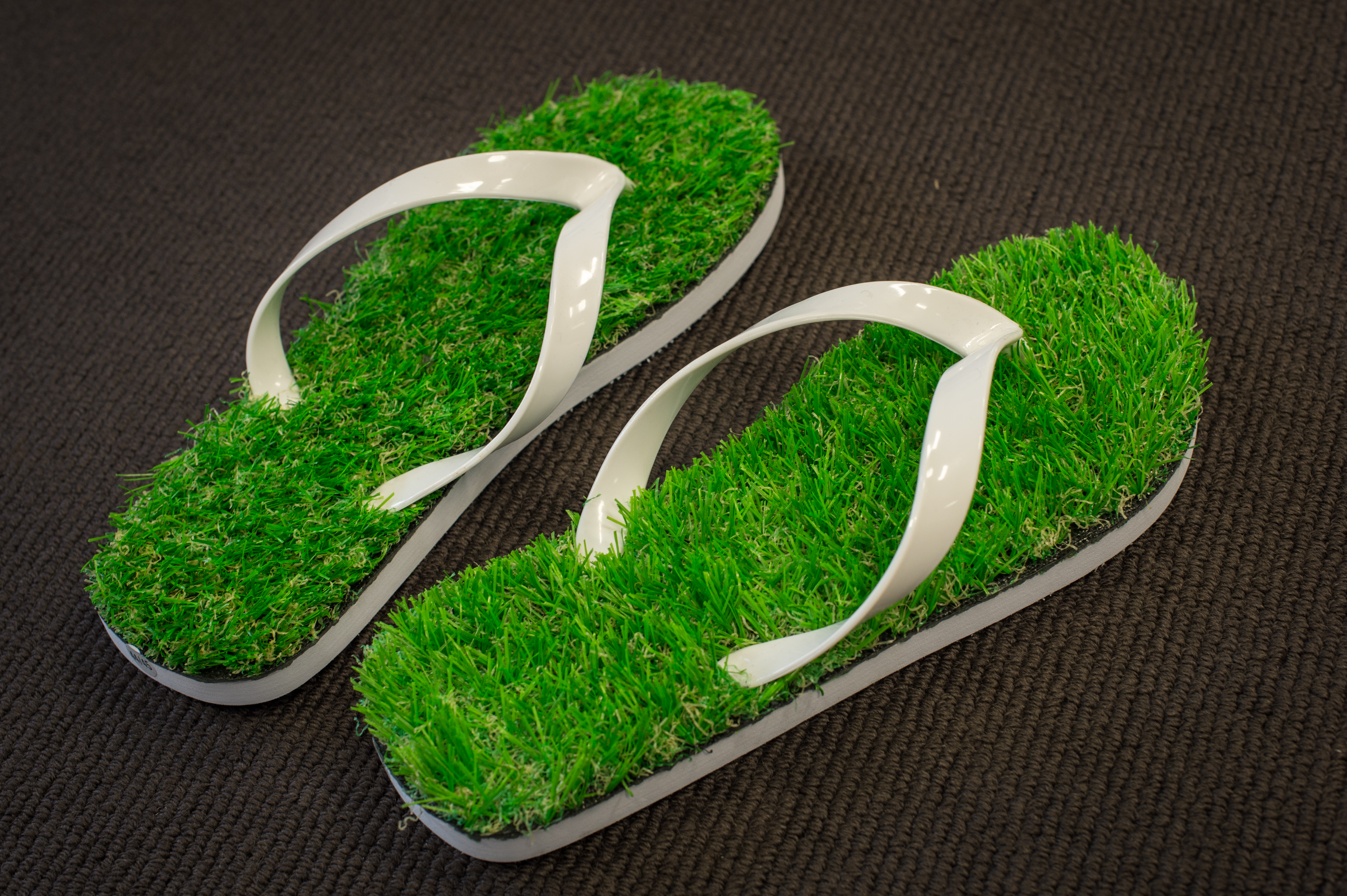 A pair of flip-flops with artificial grass soles and white straps placed on a dark surface.