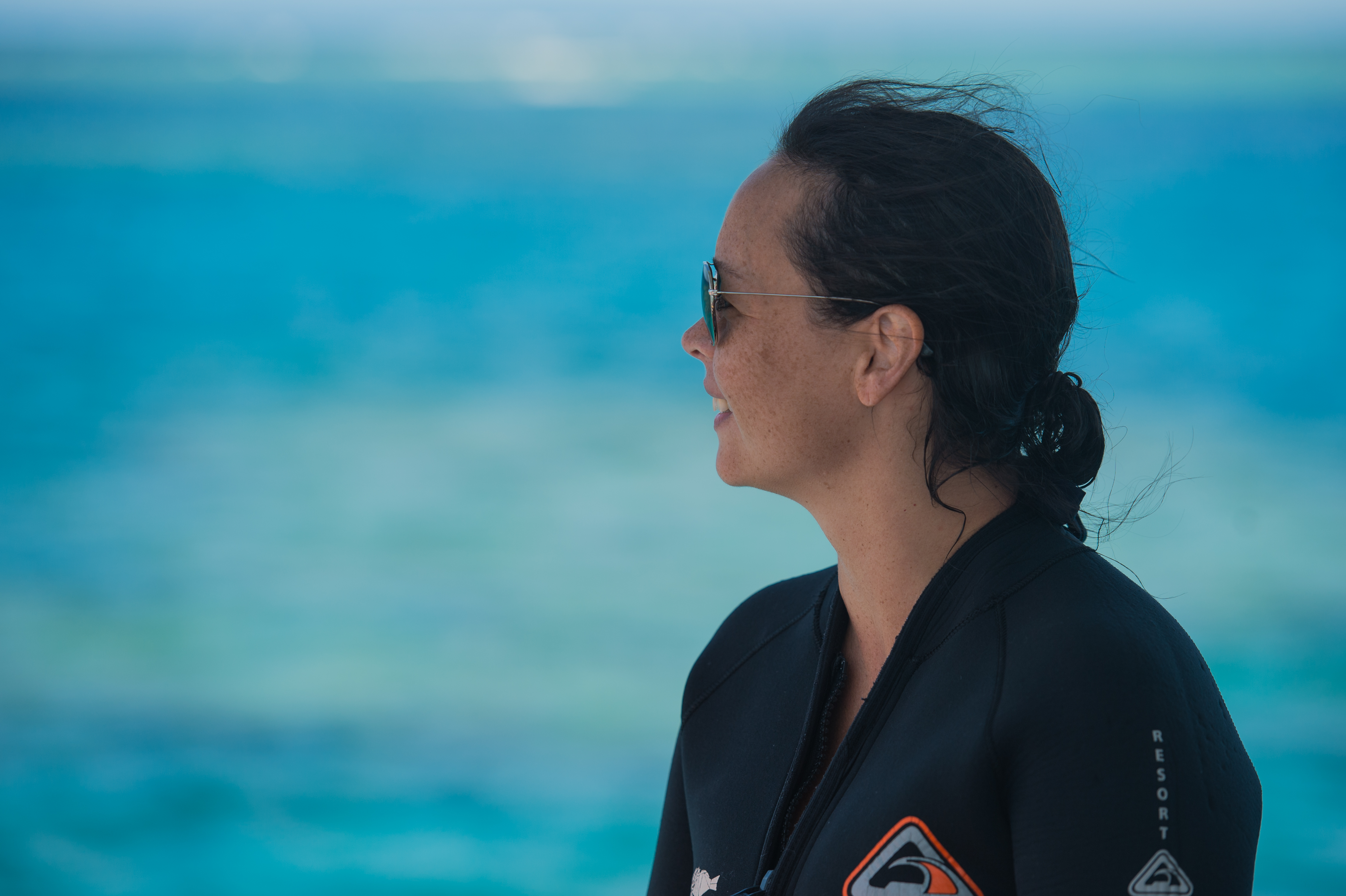 A woman wearing sunglasses and a wetsuit looks to the side with the ocean in the background.