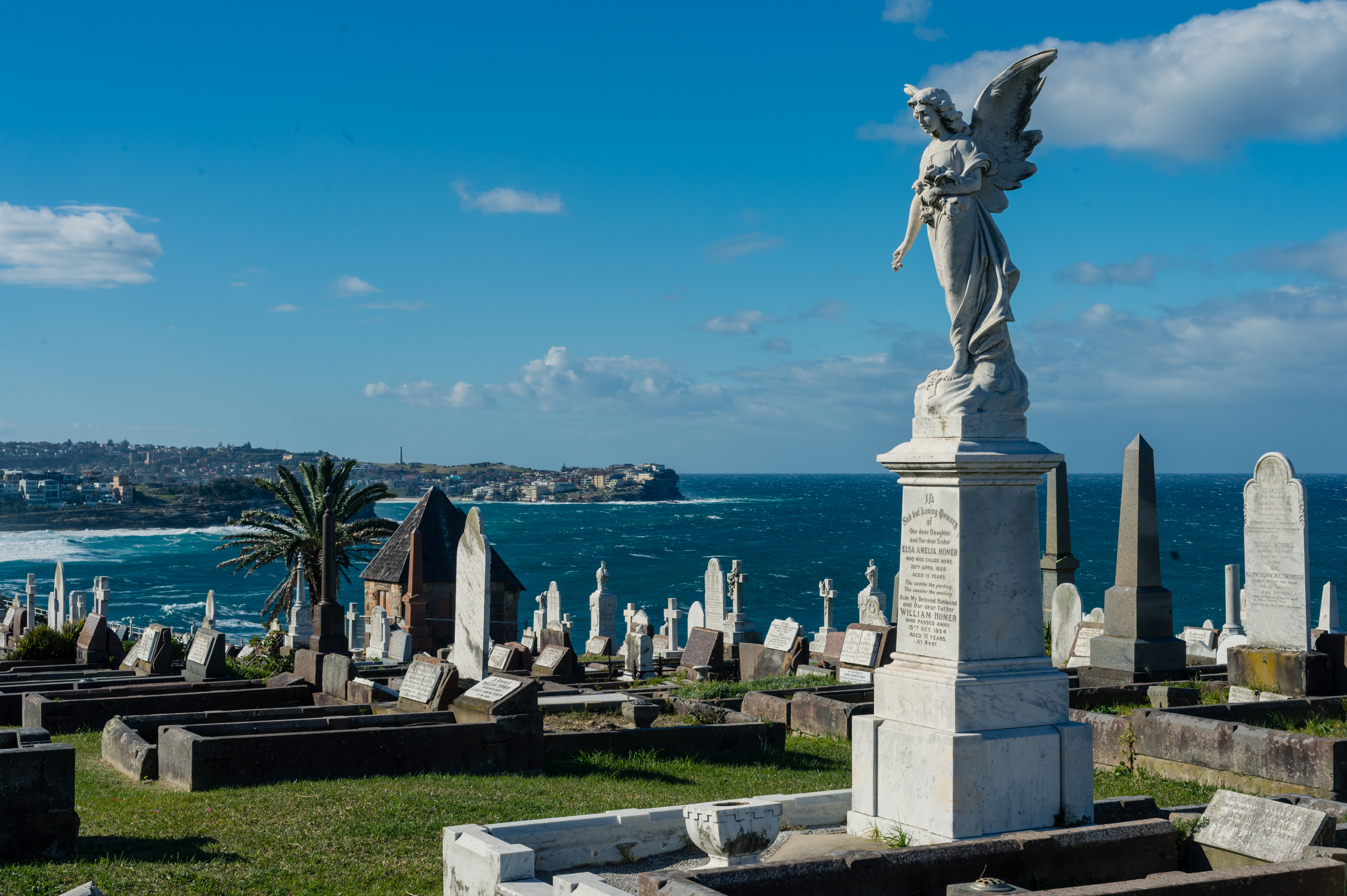A cemetery overlooking the ocean with a large angel statue on a tombstone under a clear blue sky.