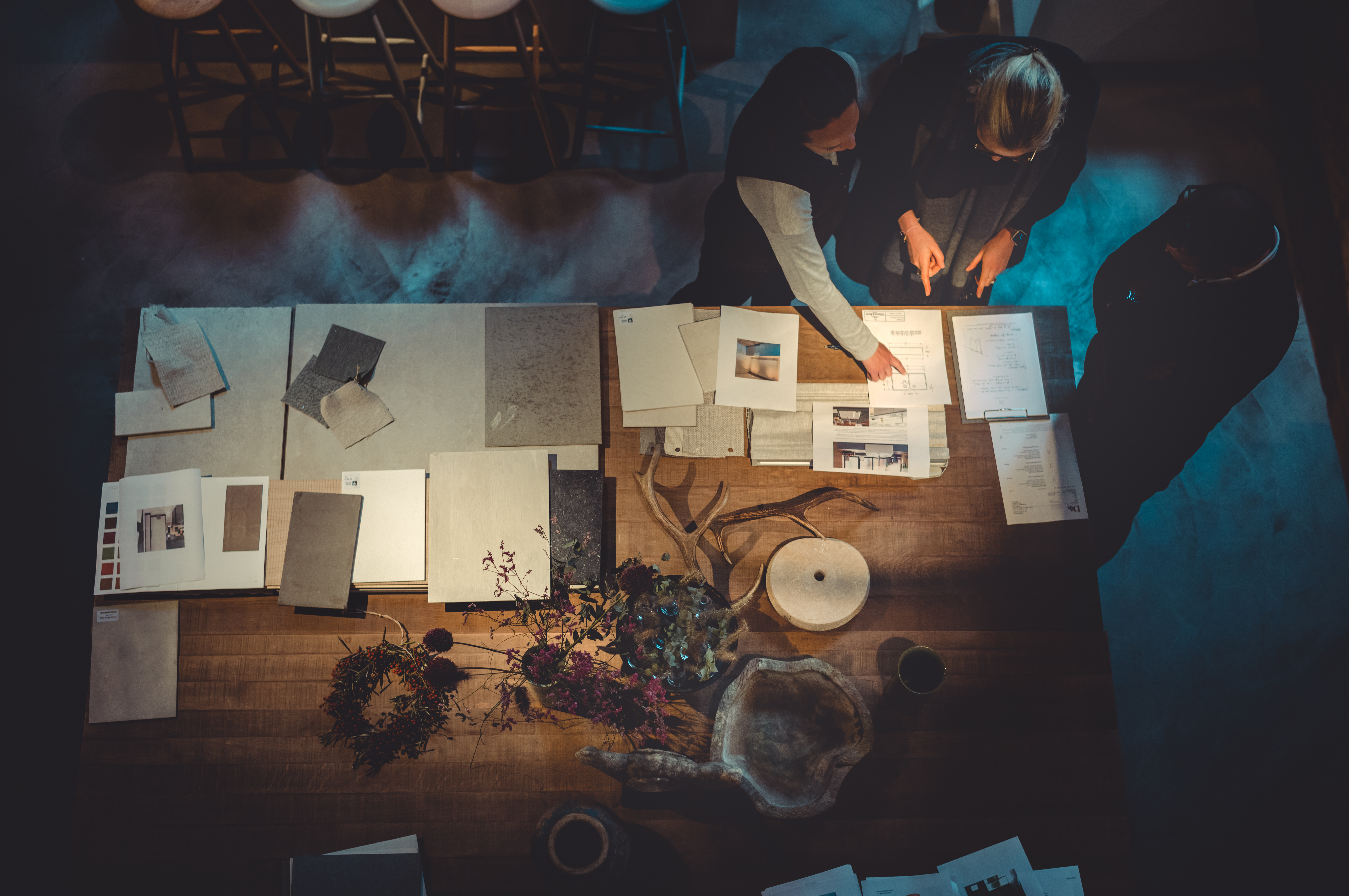 An aerial view of a wooden table, displaying various tiles, woods, stones, and fabrics in an organized manner. Three people huddle over the table, planning and designing their project.