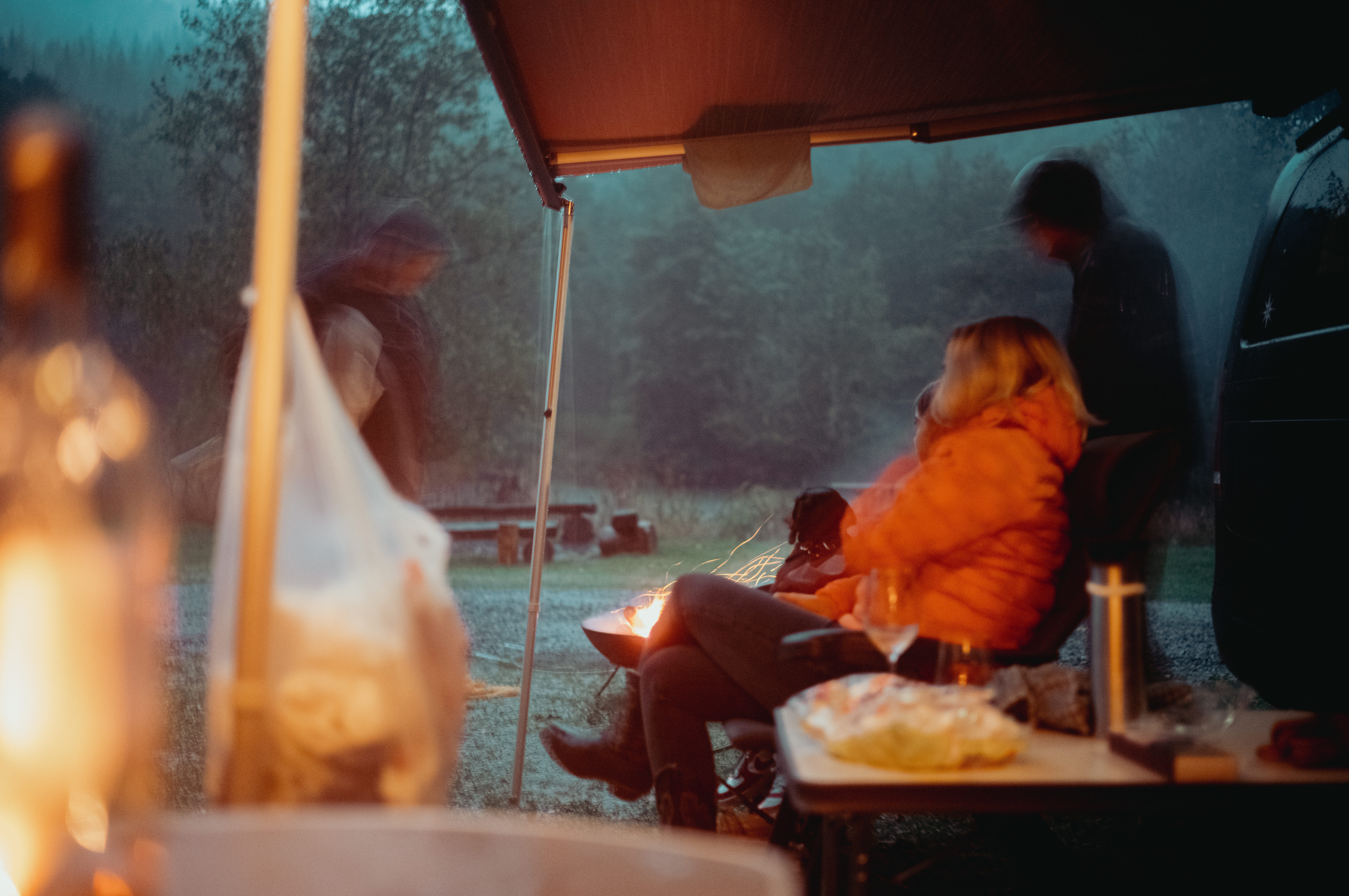 People sit under a van&#039;s awning, staying dry while a small fire burns nearby on a rainy camping trip.