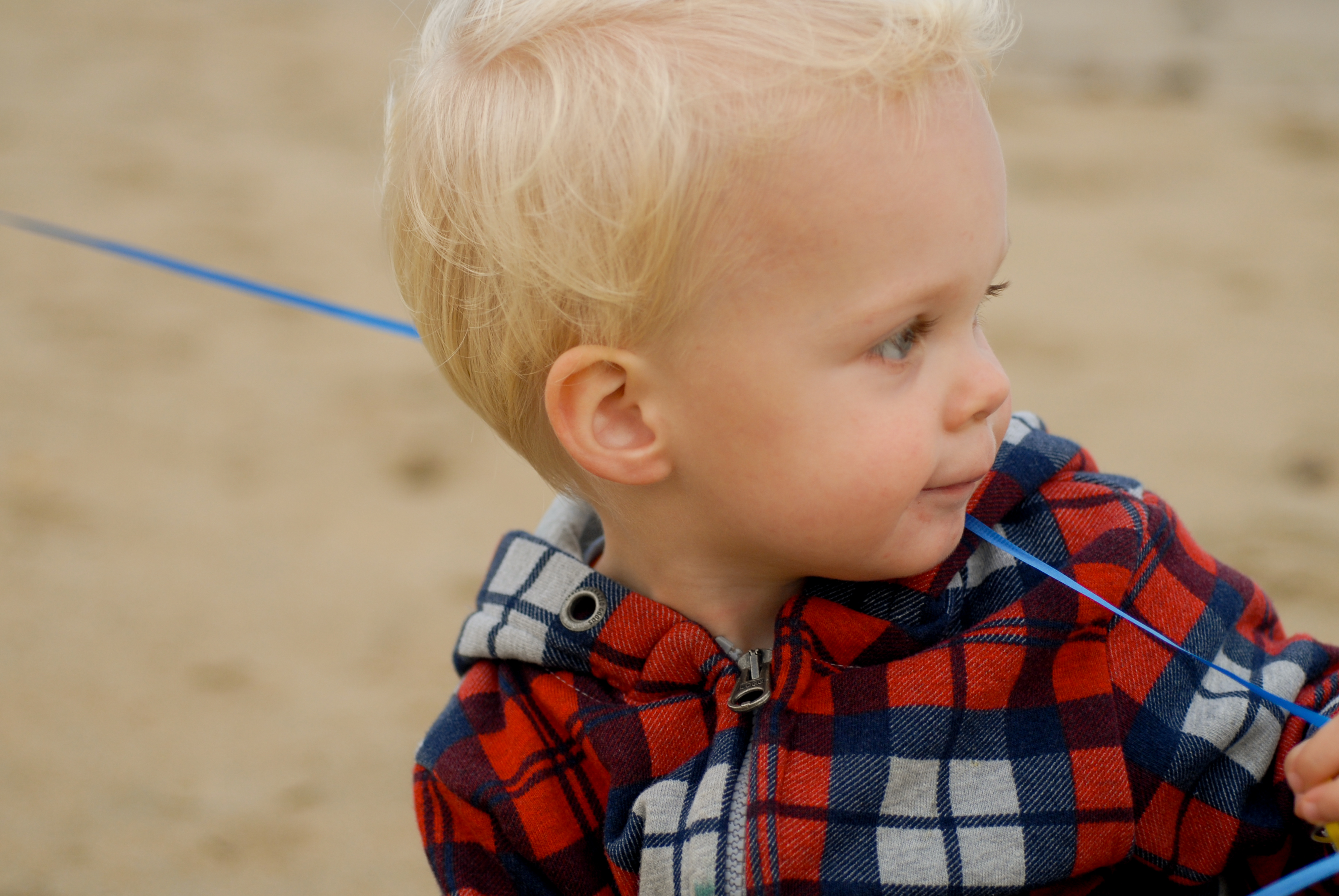 A young child in a plaid jacket holds a blue ribbon while looking to the side on a sandy beach.