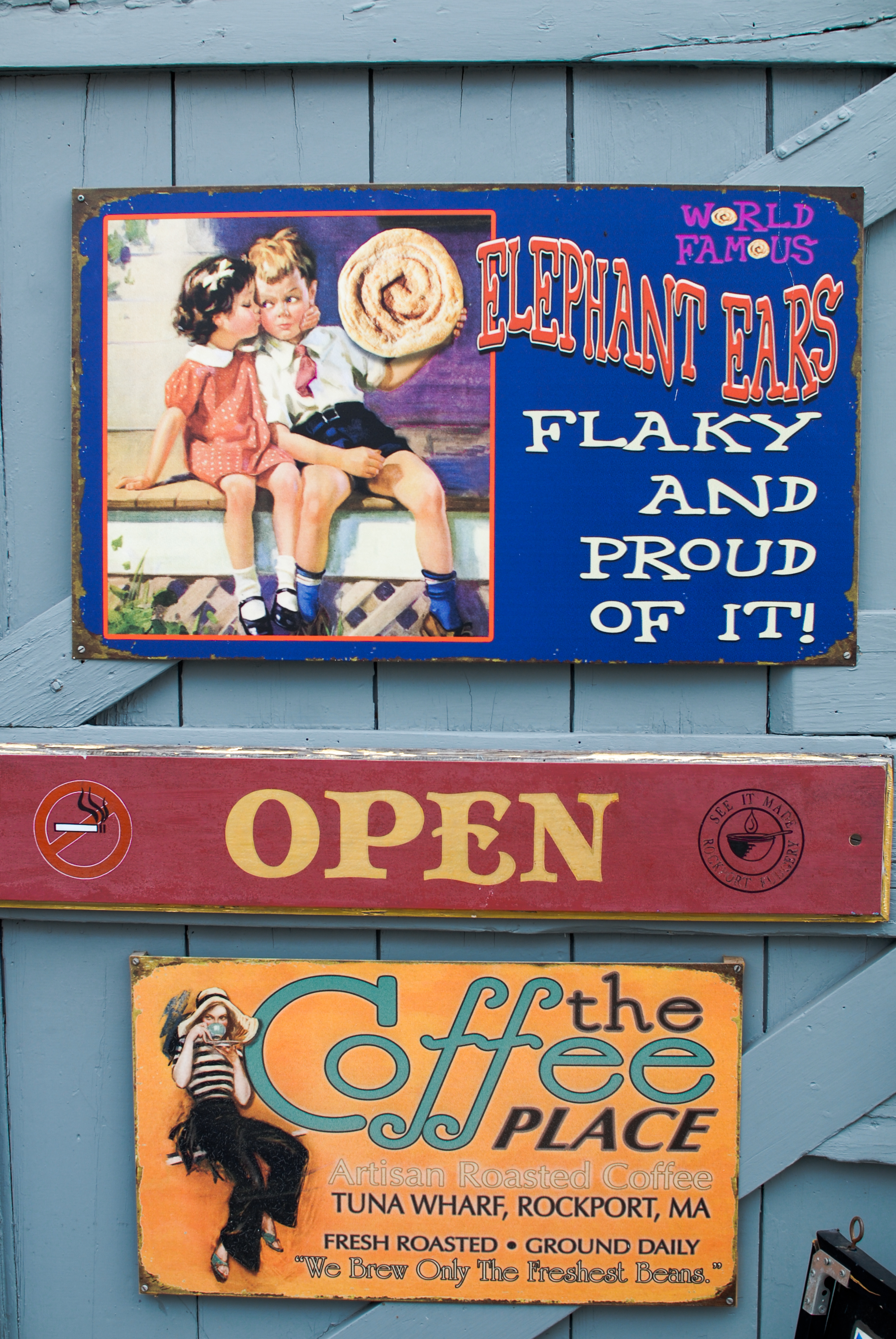 Three colorful shop signs advertising elephant ears, coffee, and an open business, mounted on a blue wooden wall.