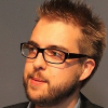 A man with glasses and a beard looks slightly to the side with a neutral expression.