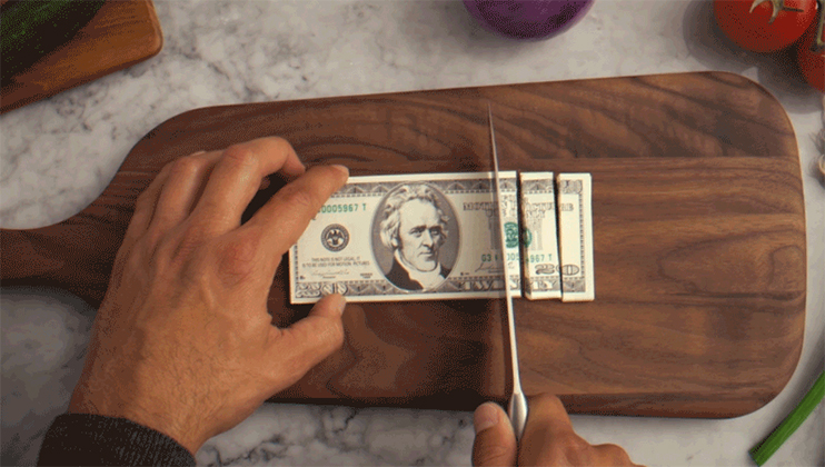 A person cuts a twenty-dollar bill into smaller pieces on a wooden cutting board using a knife.
