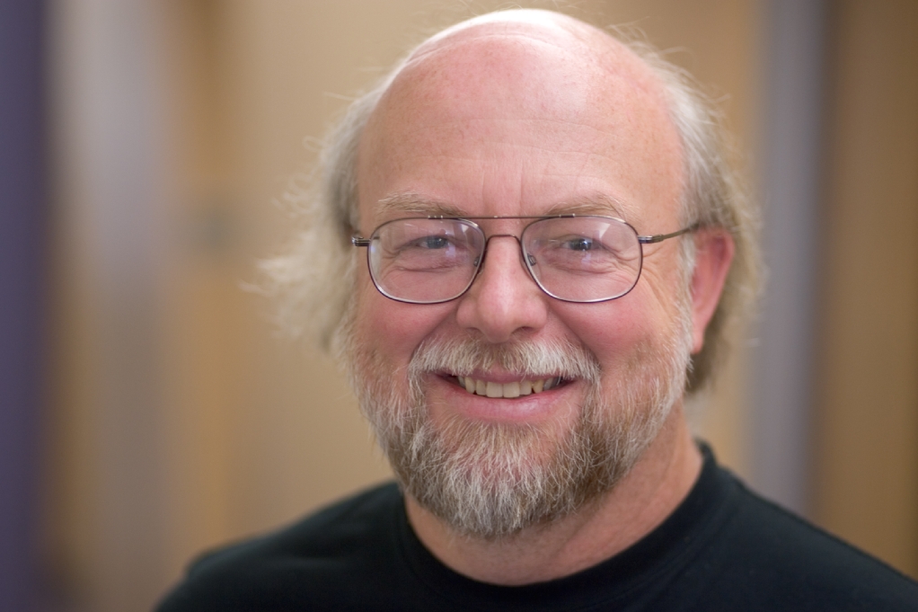 A man with glasses and a beard smiling, wearing a black shirt, in an indoor setting.