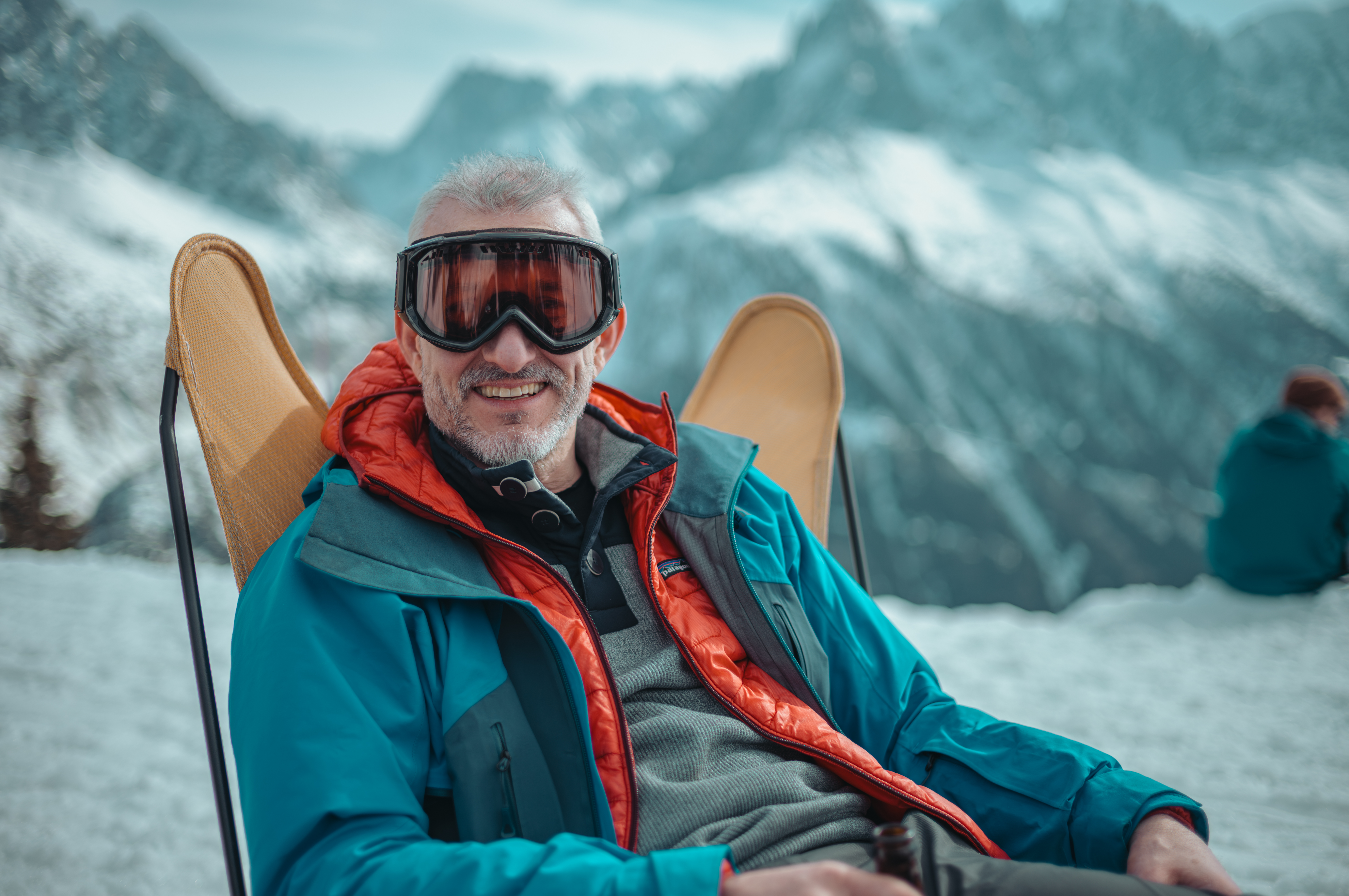 Smiling man in ski goggles relaxes in a lounge chair after skiing, holding a drink and bundled in winter layers.