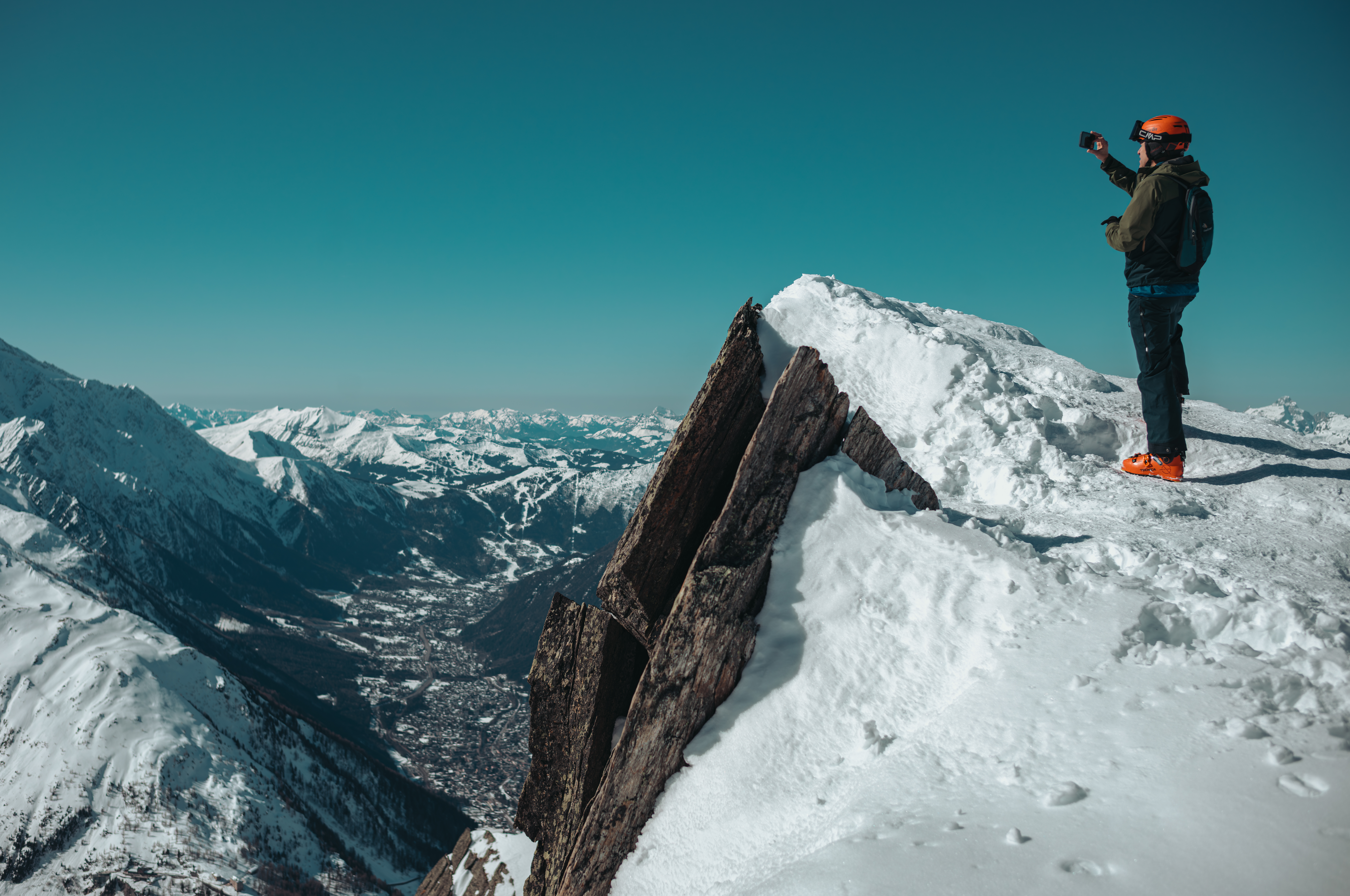 Person in orange helmet and ski boots stands on a snowy ridge at Bochard summit, photographing the Chamonix valley and surrounding snow-covered mountains.