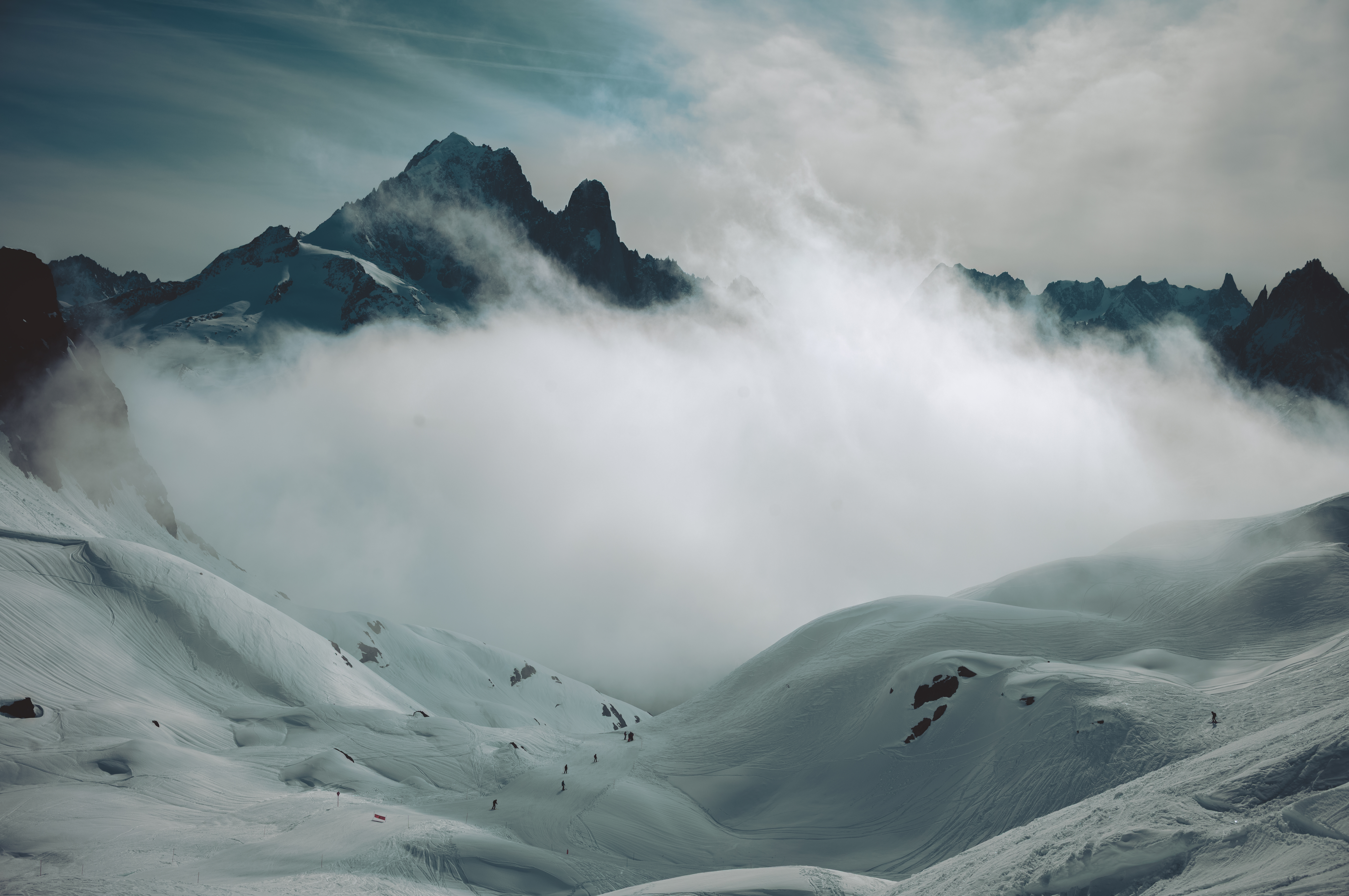 Skiers descend a wide snowy slope toward a thick bank of clouds, with mountain peaks behind.