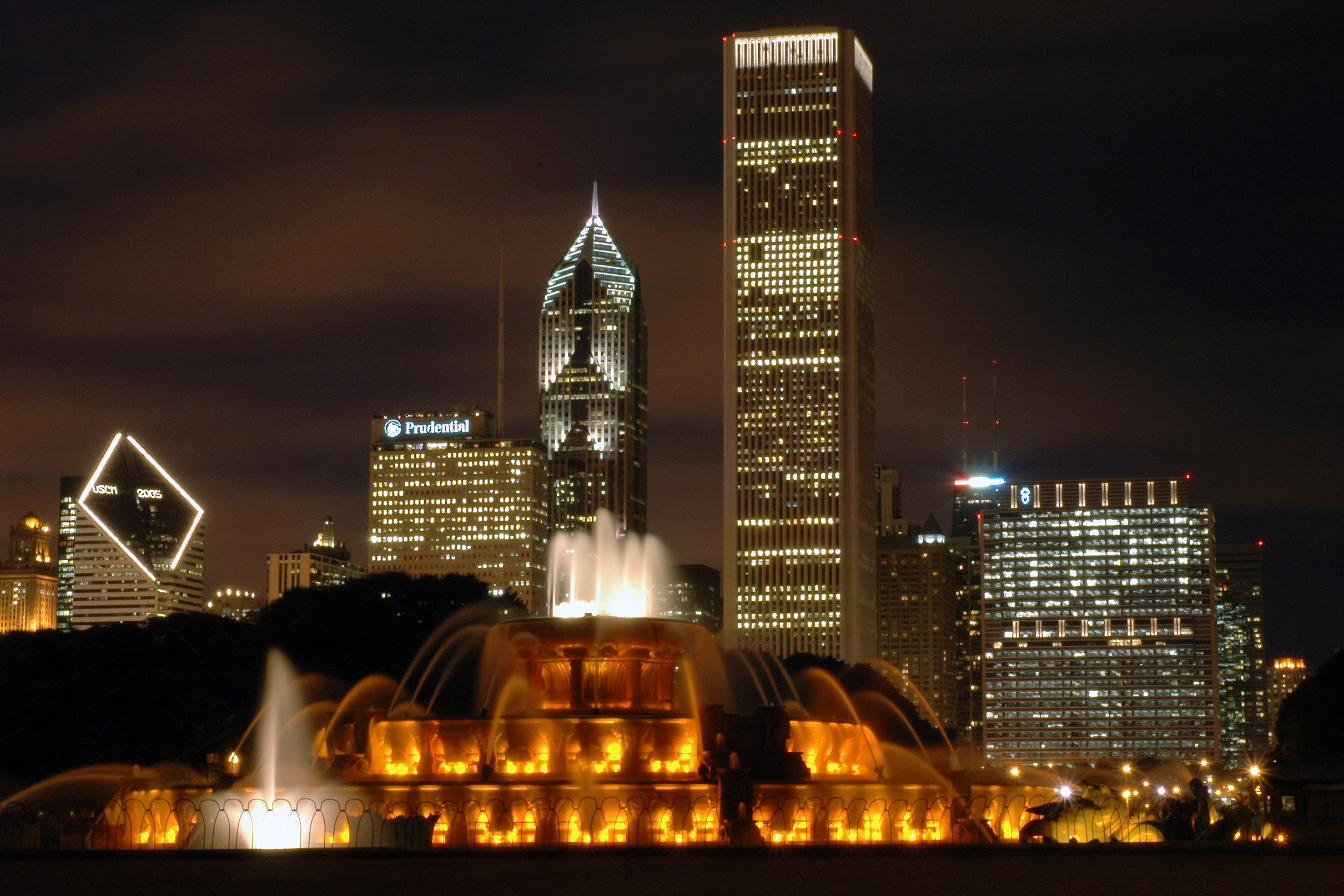 A brightly lit fountain with water jets in front of Chicago's illuminated skyline at night.