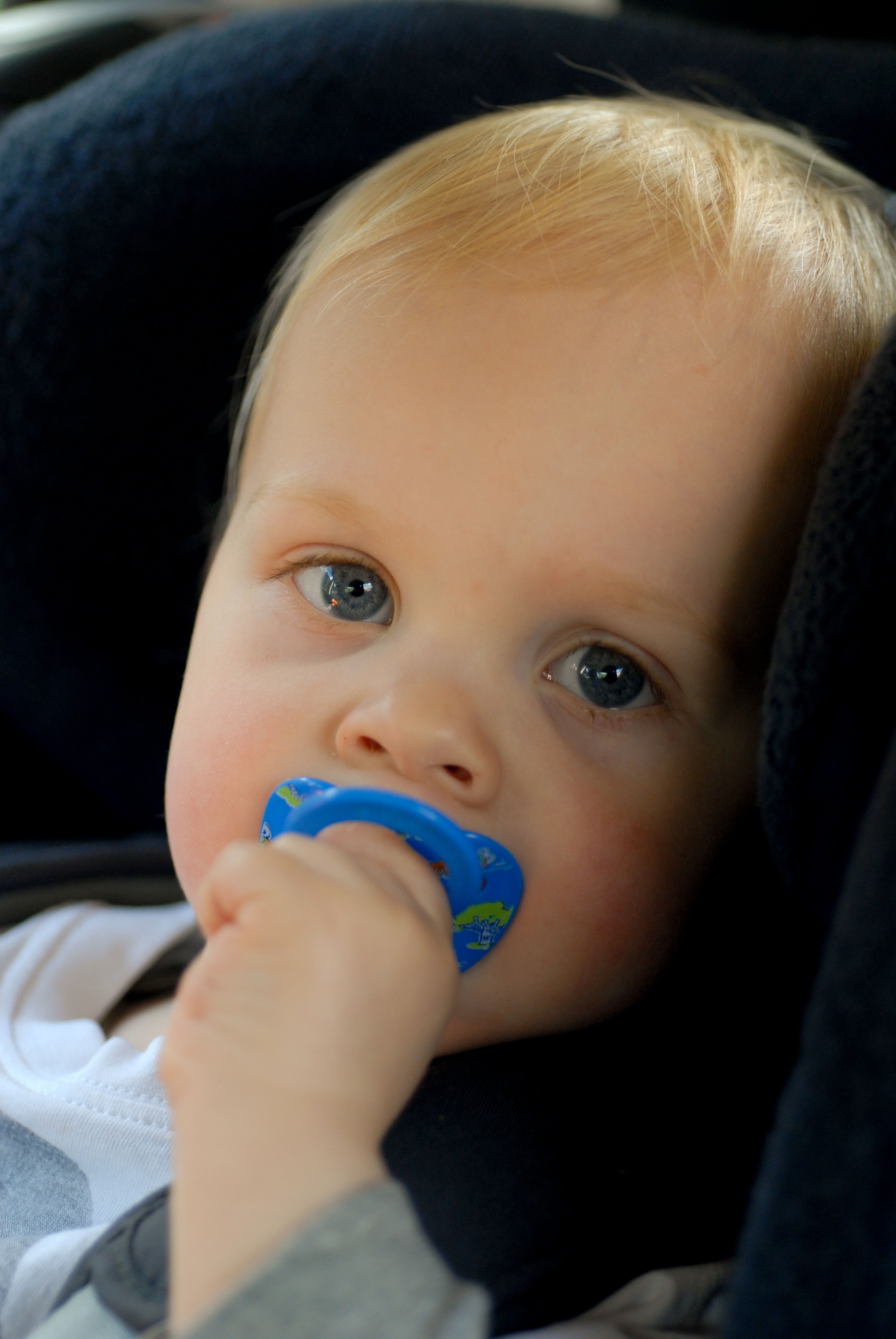 A baby with a blue pacifier sits in a car seat, looking ahead.