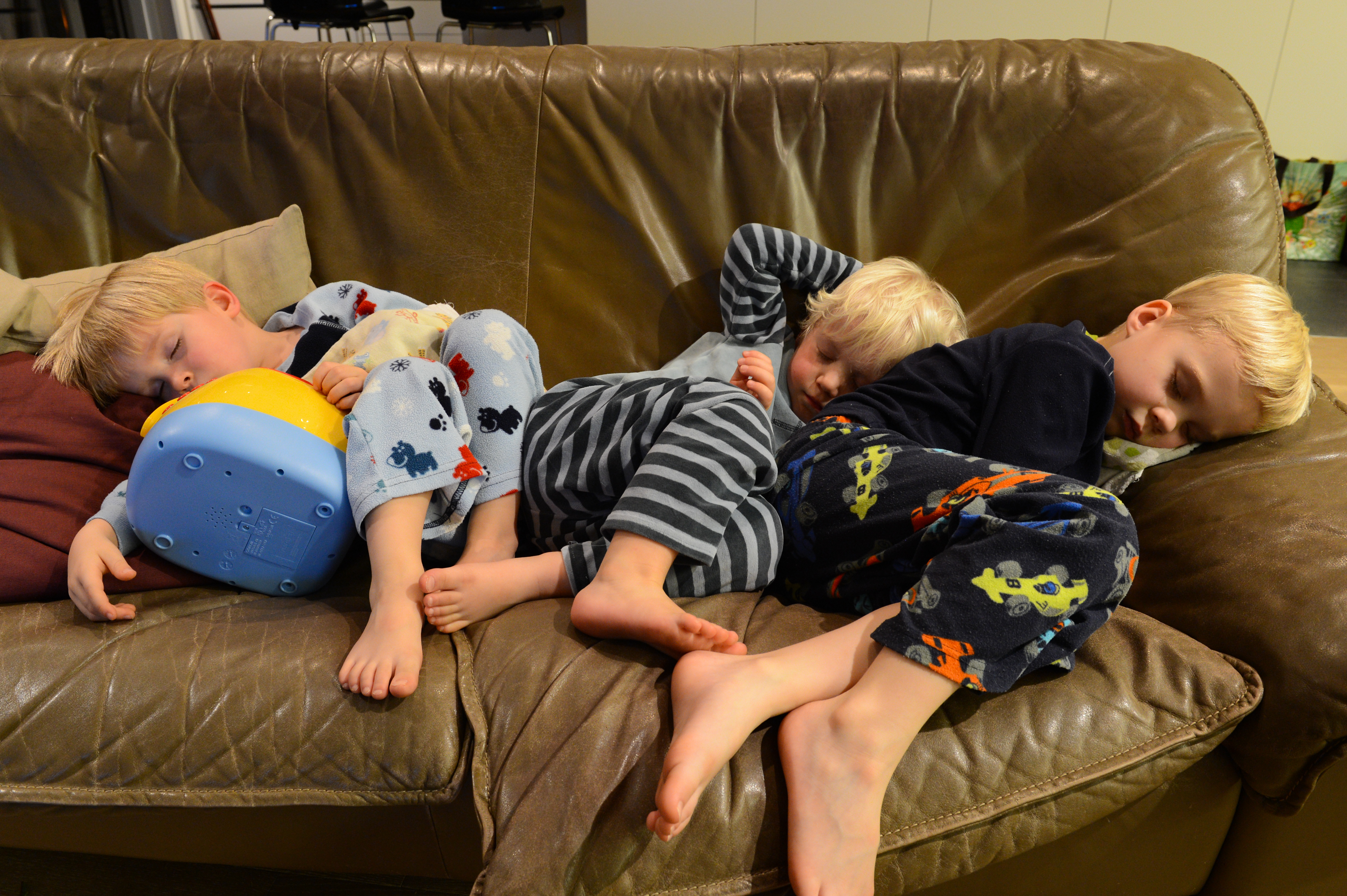 Three young children in pajamas are asleep on a brown couch, curled up closely together after playing.