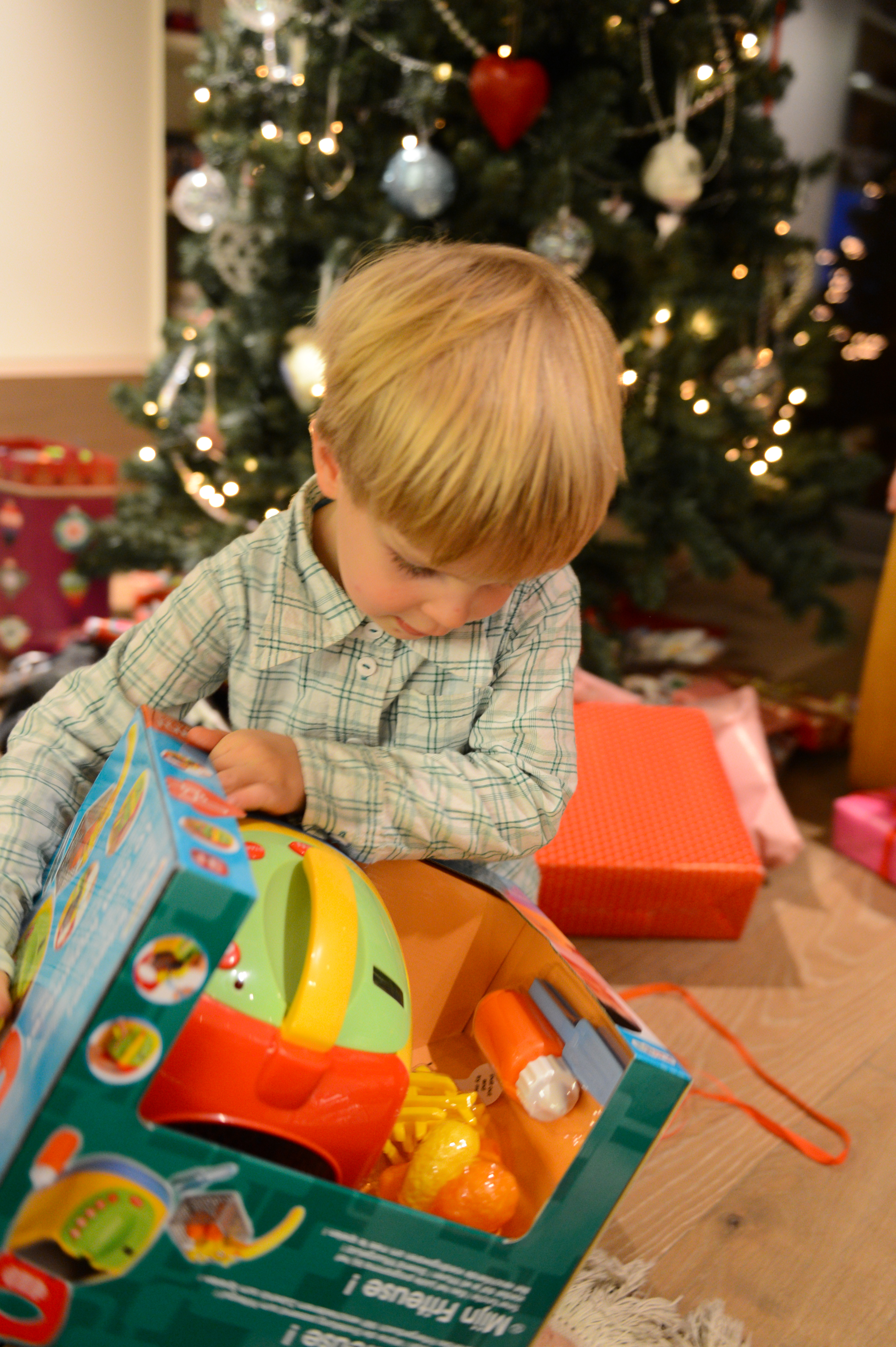 A young child opens a toy gift near a decorated Christmas tree with lights and ornaments.
