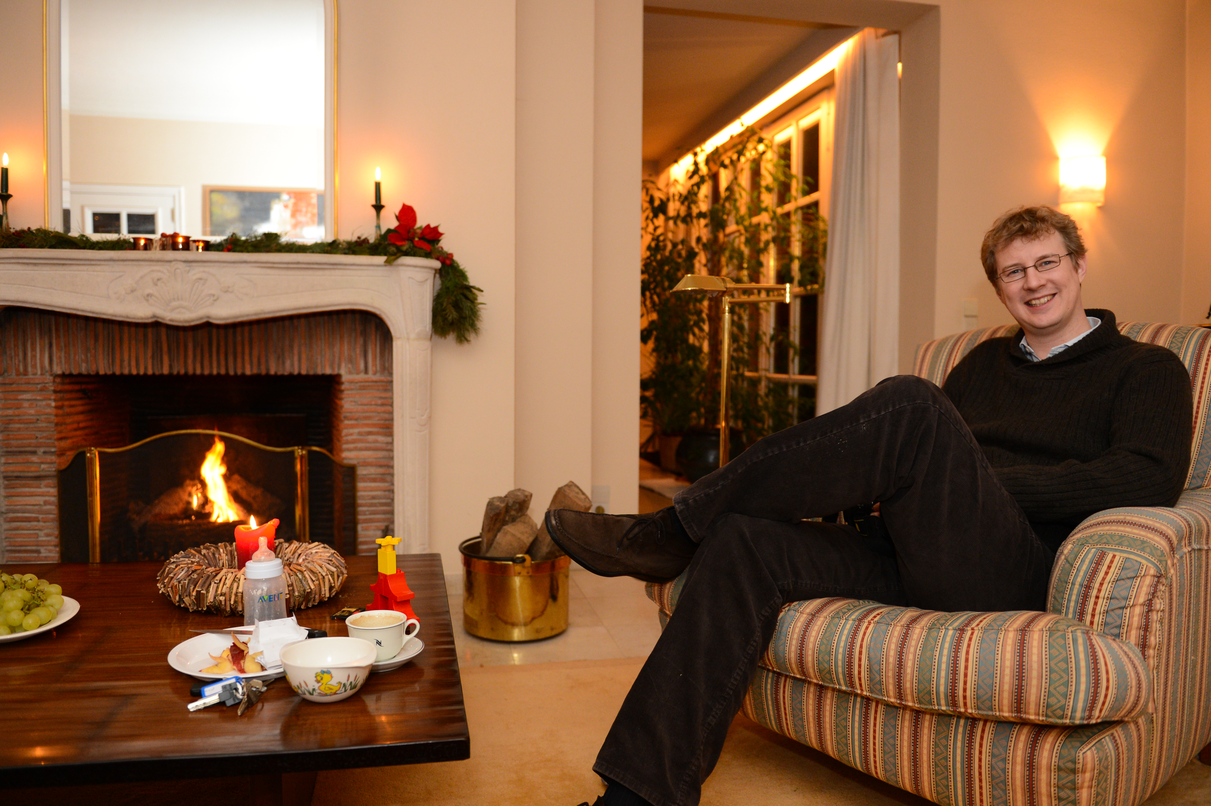 A man sits in a striped armchair, smiling, near a fireplace with holiday decorations and a table with food.