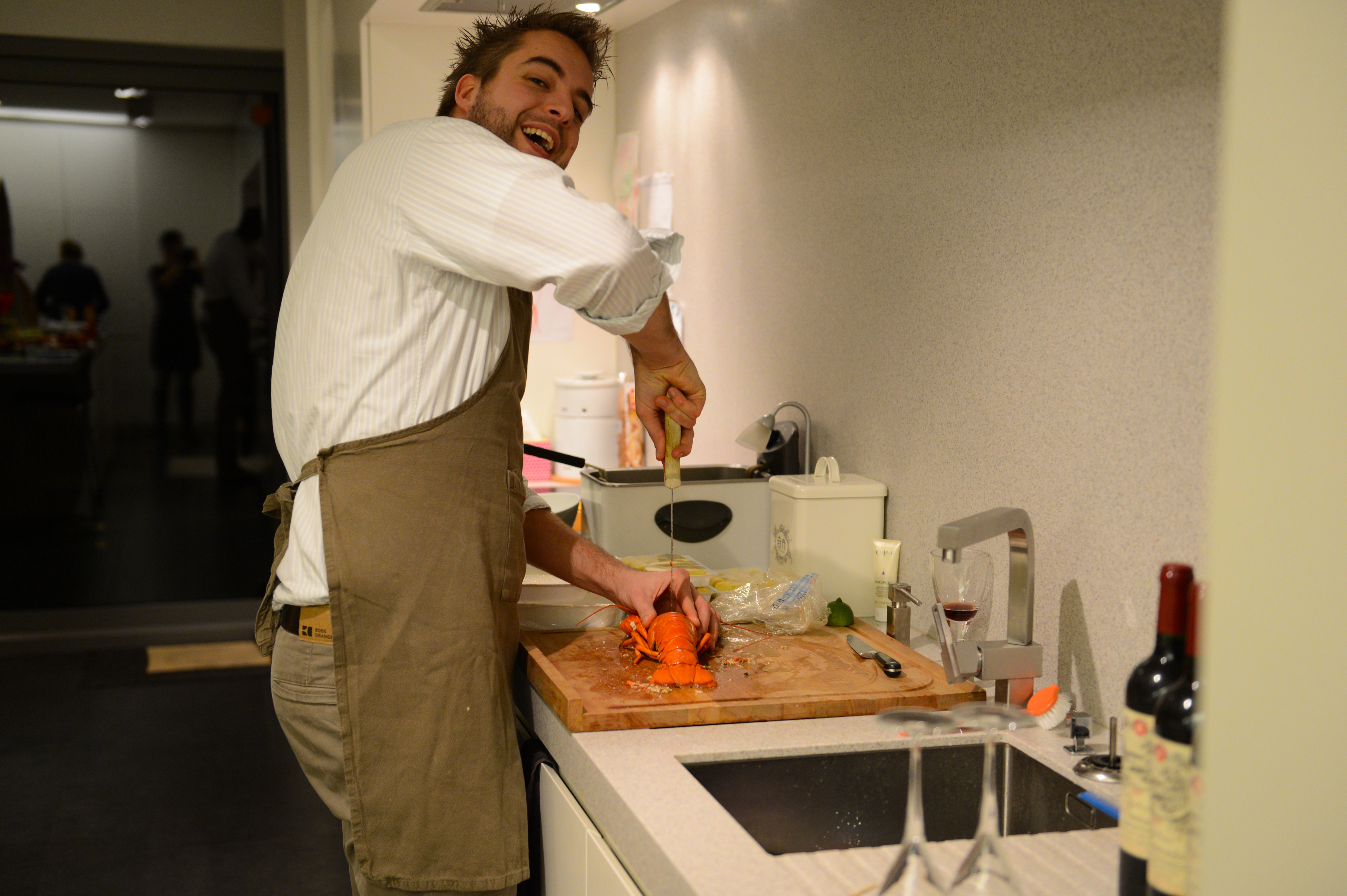 A man in an apron smiles while preparing a lobster on a cutting board in a kitchen.