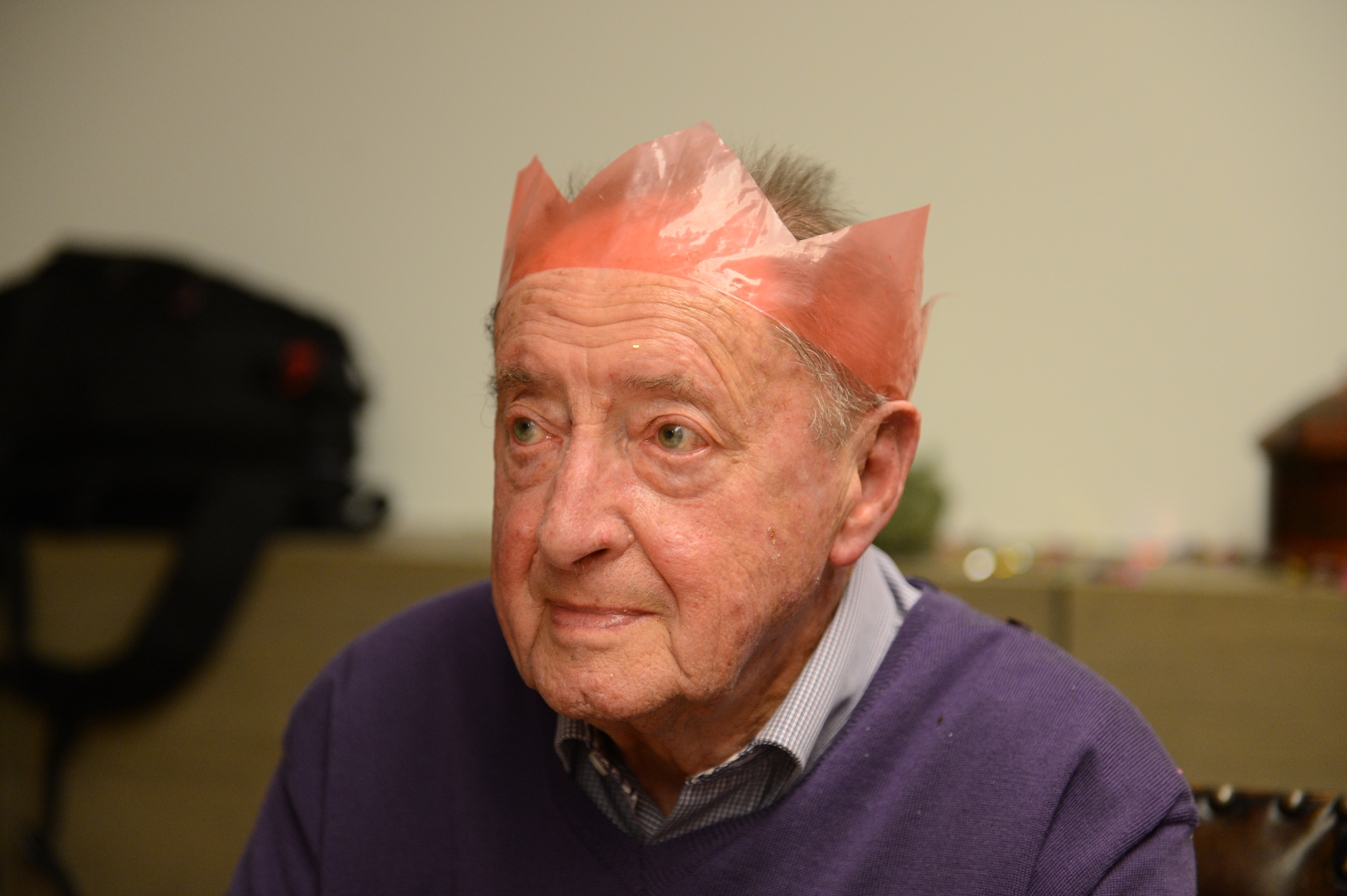 Elderly man wearing a red paper crown sits indoors, looking thoughtful during a Christmas Eve gathering in Ekeren.
