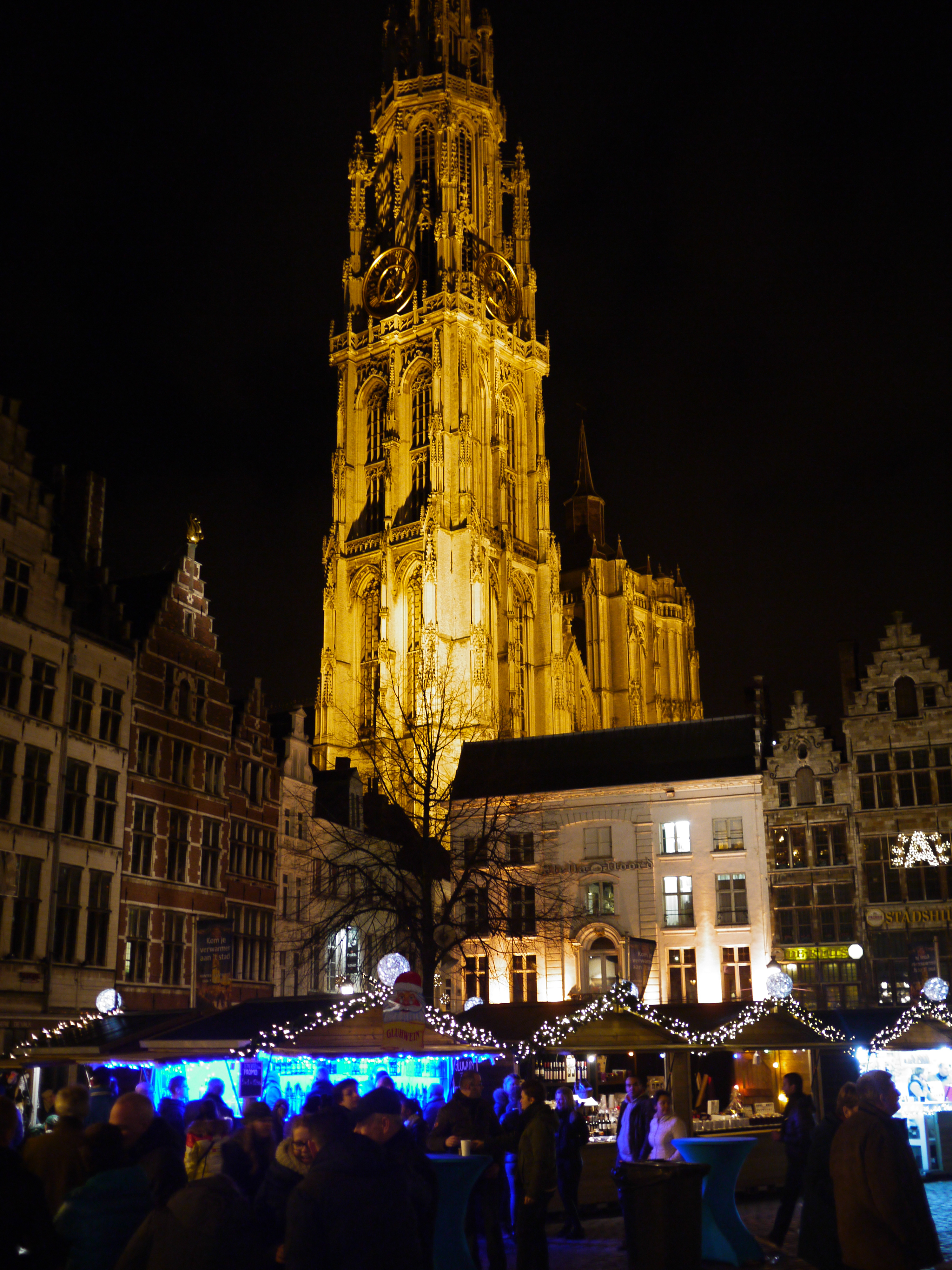 People gather at a Christmas market in Antwerp, with festive stalls and a brightly lit church tower in the background.