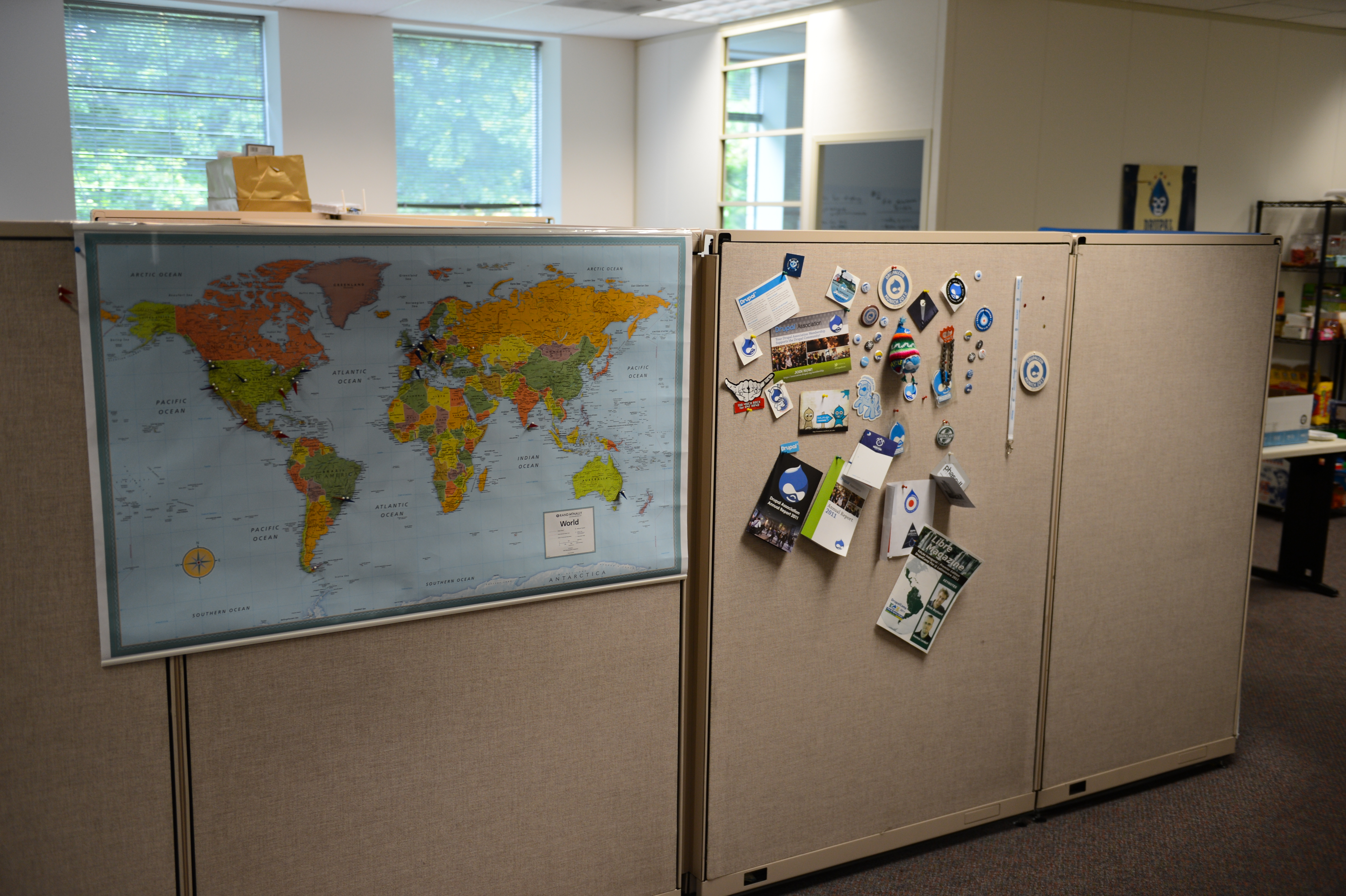 Office cubicle with a large world map and a bulletin board covered in Drupal-related pins, stickers, and notes.