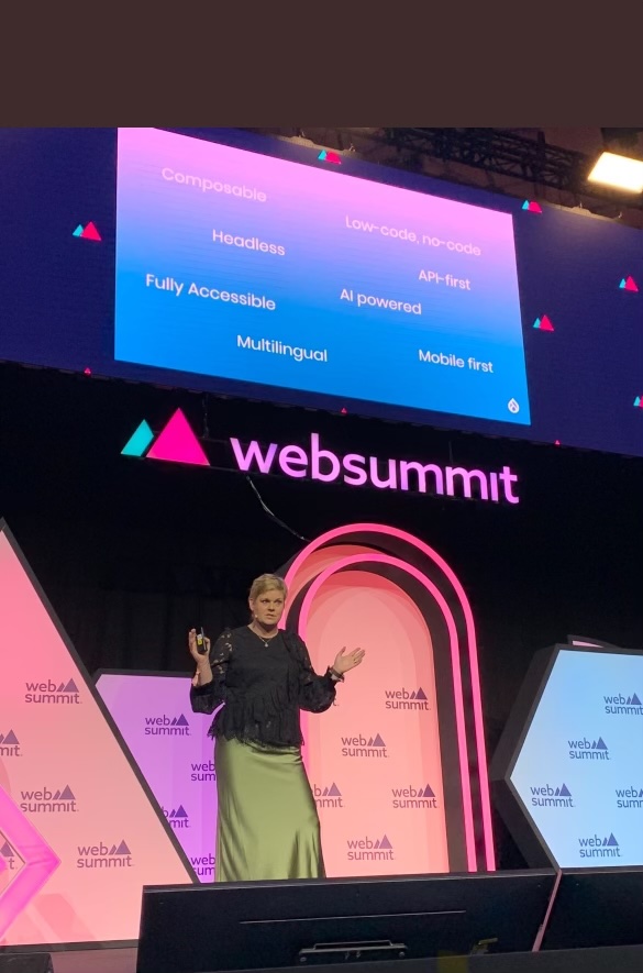 A presenter at Web Summit with a screen behind her listing tech terms like 'Composable', 'Headless', 'AI powered', 'multi-lingual' and more.