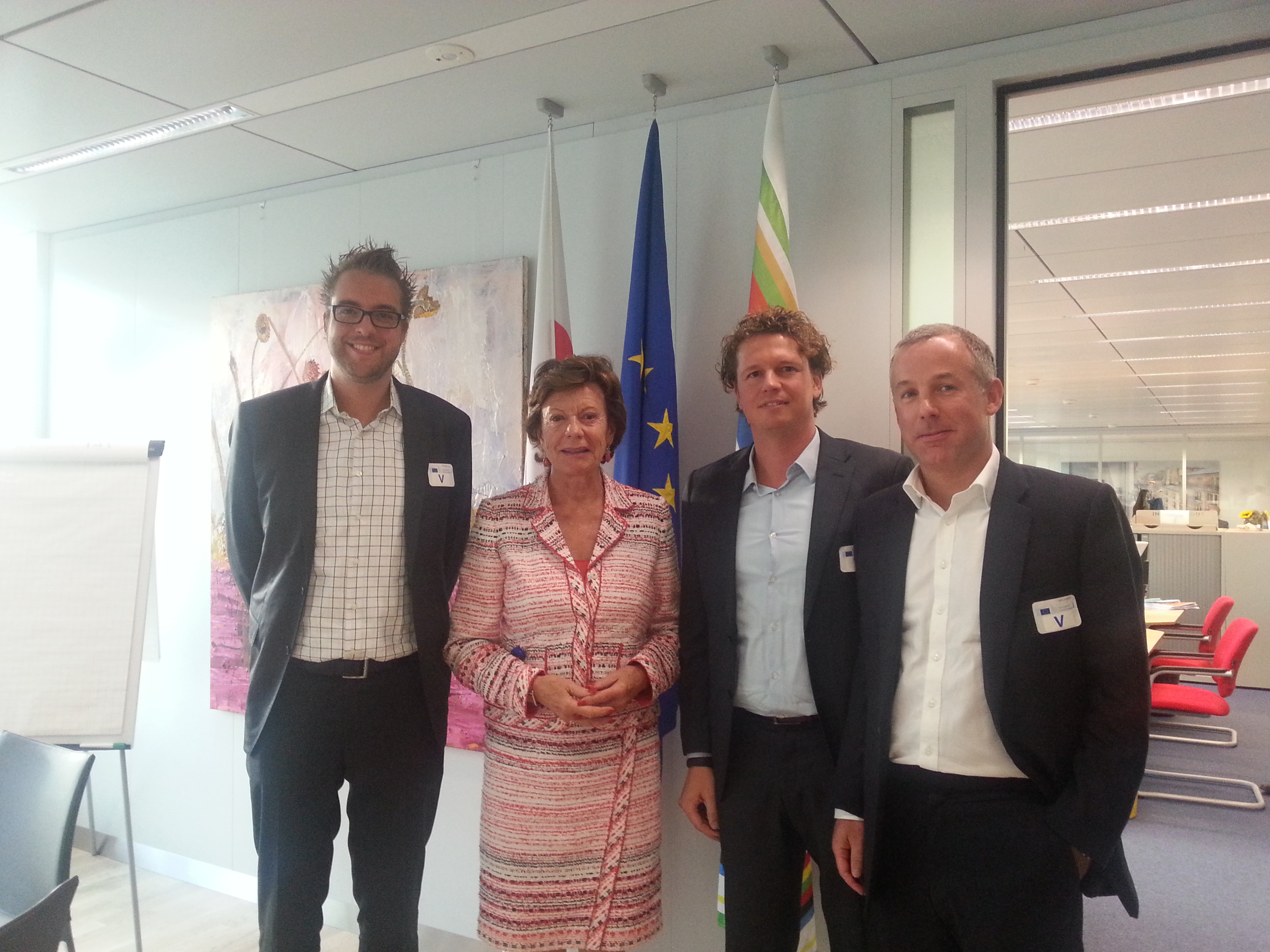 Four people stand together in an office, posing for a group photo in front of flags.