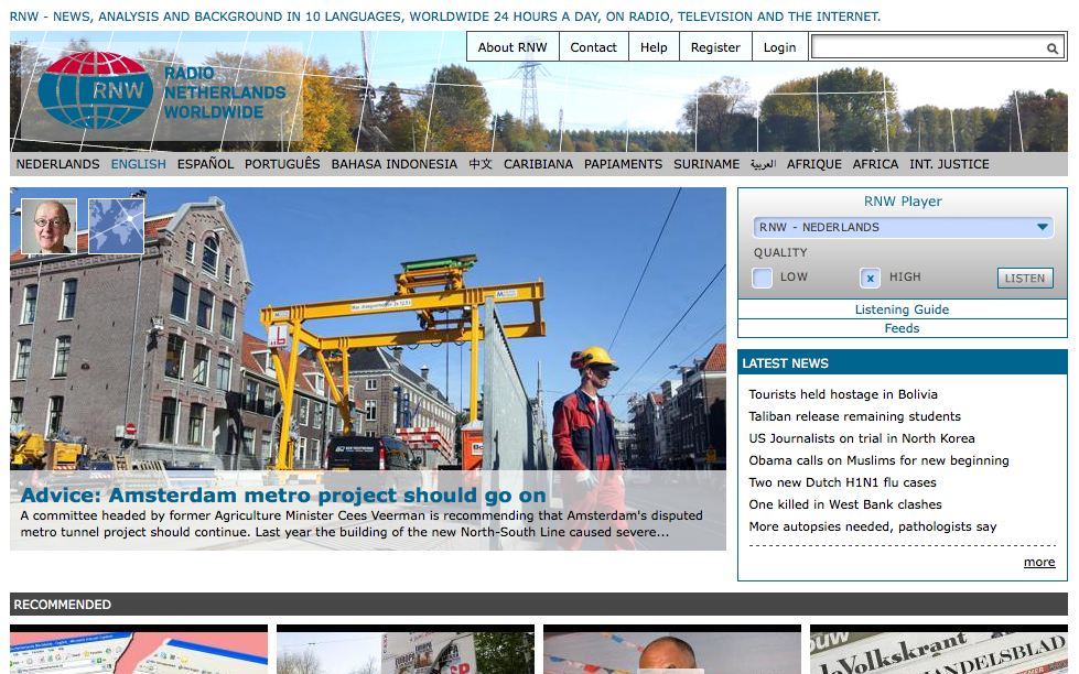 A construction worker in a yellow helmet stands near a large yellow crane at an Amsterdam metro project site.