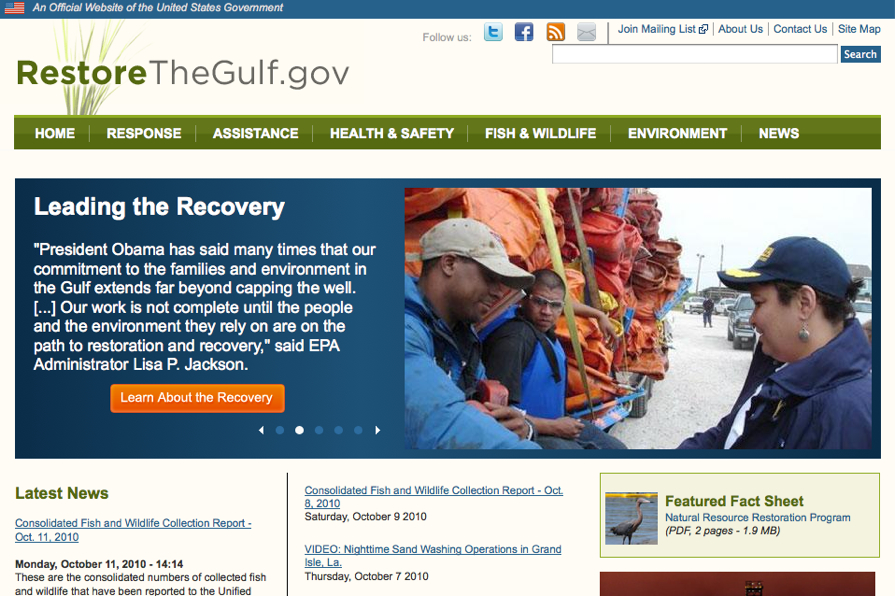 A government official speaks with workers in front of stacked orange equipment, discussing Gulf Coast restoration efforts.