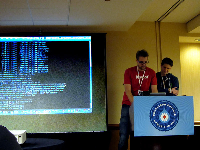 Two men on stage, standing behind a laptop on a pedestal, typing Git commands as they are displayed on a large screen behind them.