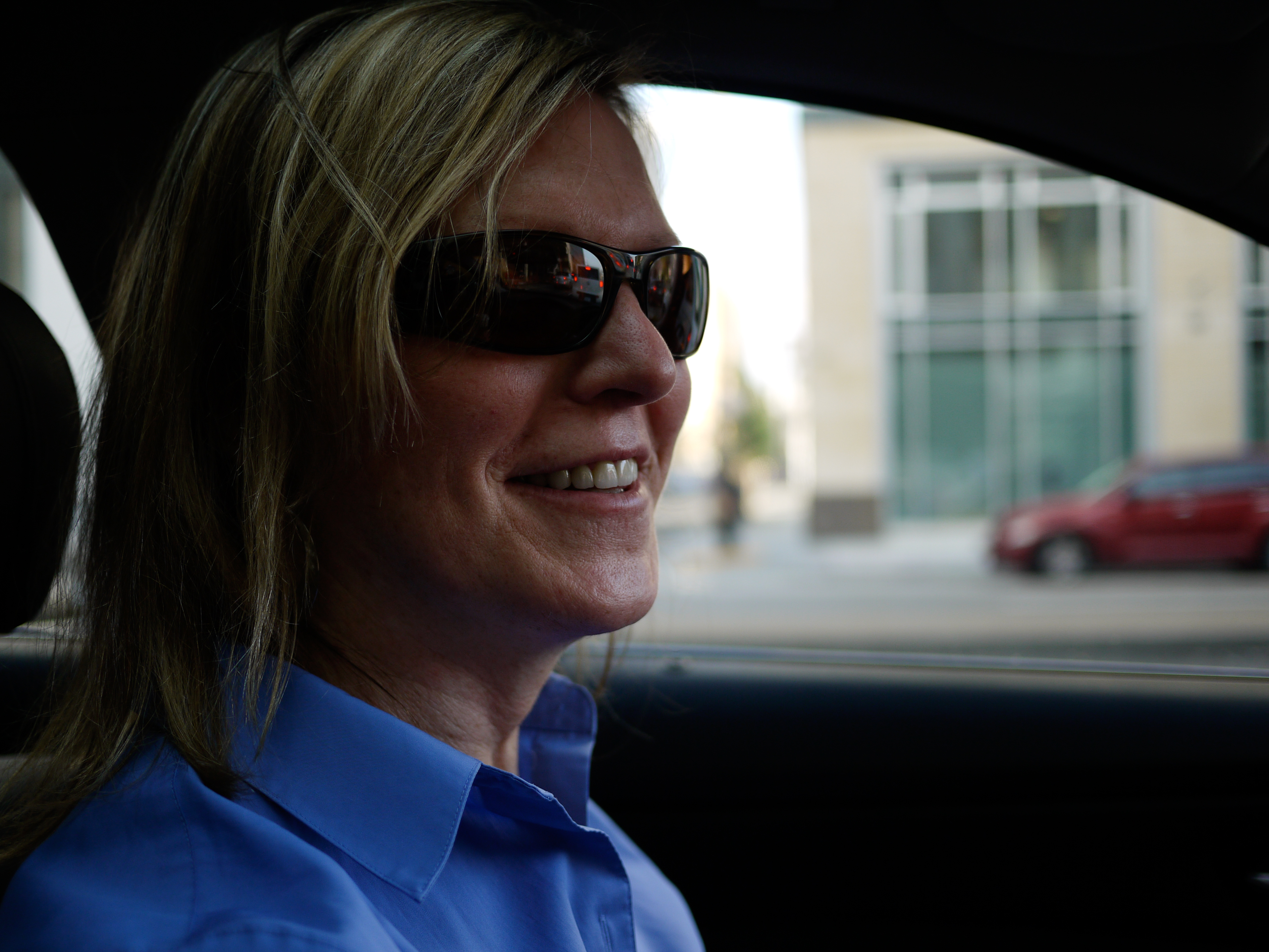 A person wearing sunglasses and a blue shirt smiles while sitting in a car.