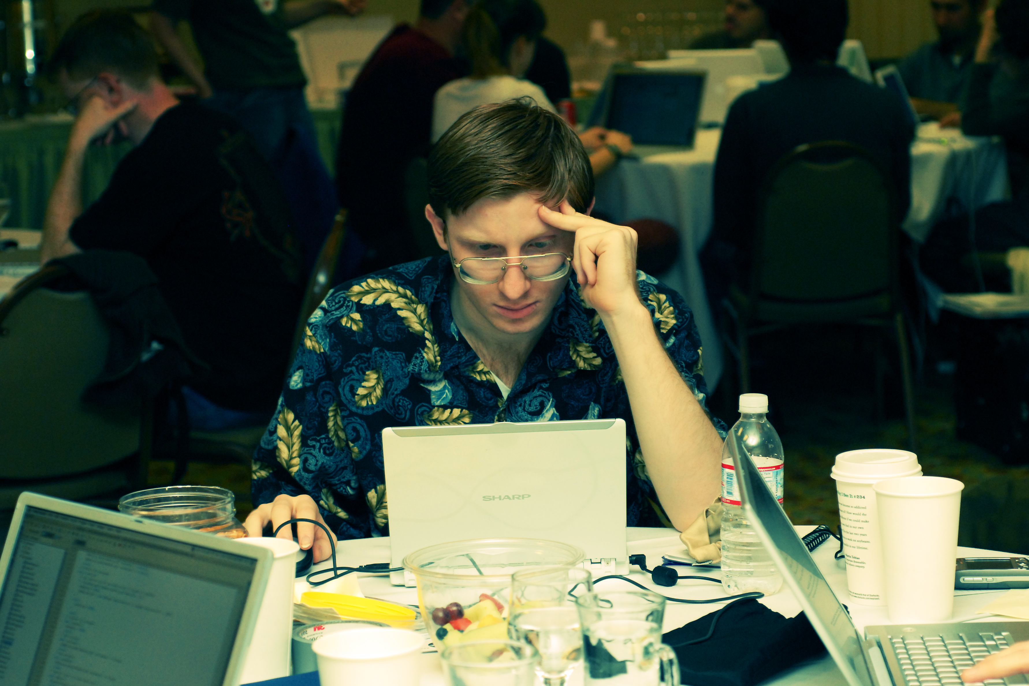 A man in a patterned shirt focuses intently on his laptop at a conference table with drinks and laptops.